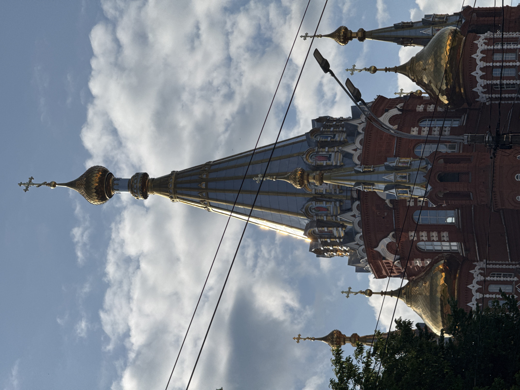 The image shows the Holy Trinity Cathedral in Izhevsk, Russia, on a partly cloudy day. The cathedral is a prominent red brick building with multiple golden domes and spires topped with crosses. The main spire is tall and conical, clad in silver-colored metal with gold trim. Several smaller golden domes are visible, some partially obscured by trees on the left. Overhead, several power lines stretch across the frame, and a street lamp is positioned on the right side. The sky is a mix of blue and white clouds, with sunlight reflecting off the metallic surfaces of the cathedral.