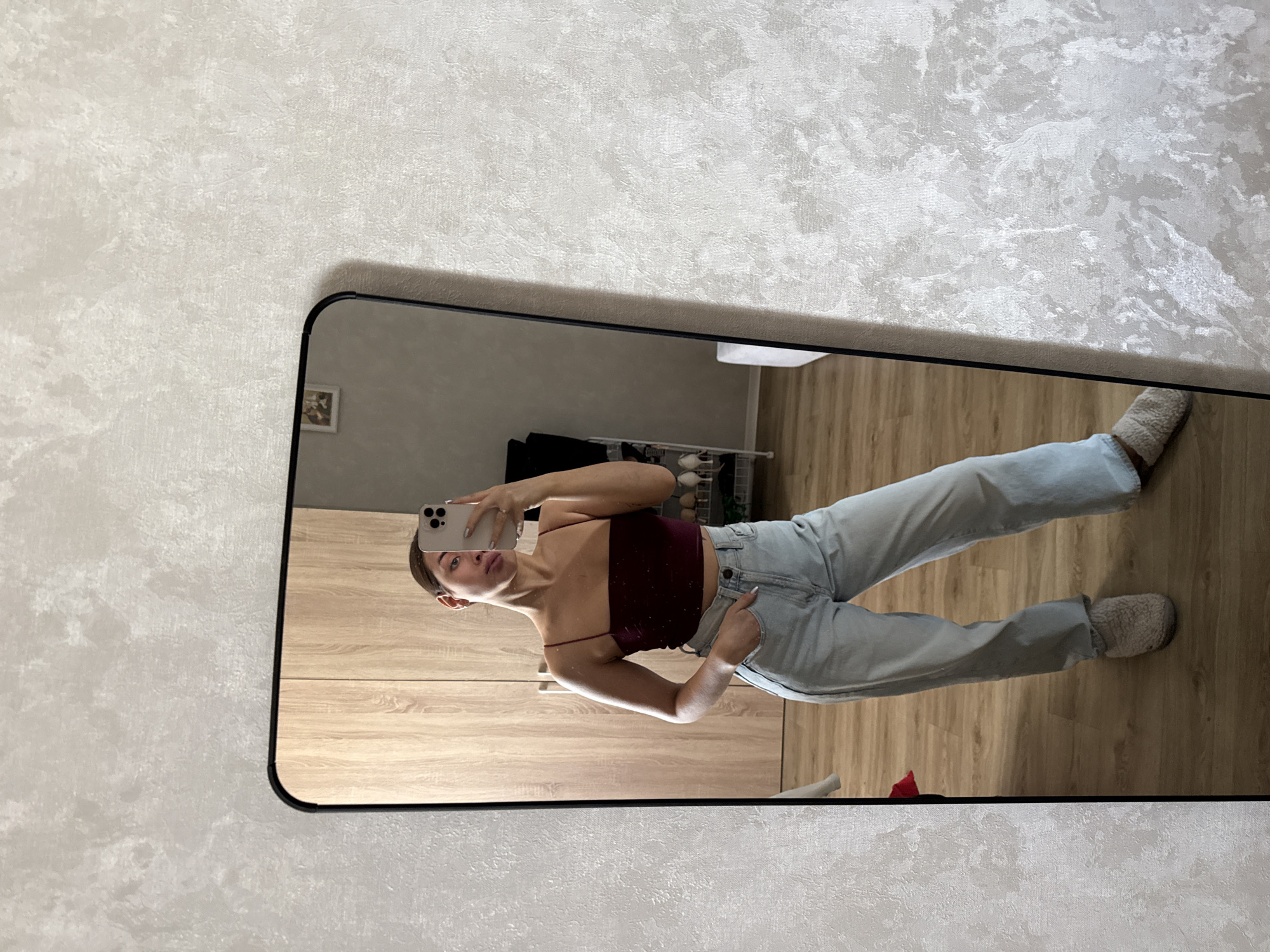 A person takes a mirror selfie indoors. The reflection reveals a light-skinned individual with dark hair pulled back, wearing a maroon camisole and light blue jeans with hands in their pockets. They are holding a silver iPhone. The background visible in the mirror includes wooden flooring, a light wooden wardrobe, a black piece of furniture, and a white shelf with items. The mirror is rectangular with a black frame, hanging on a wall covered with light-colored textured wallpaper. The location is likely in Лобаново, Russia.
