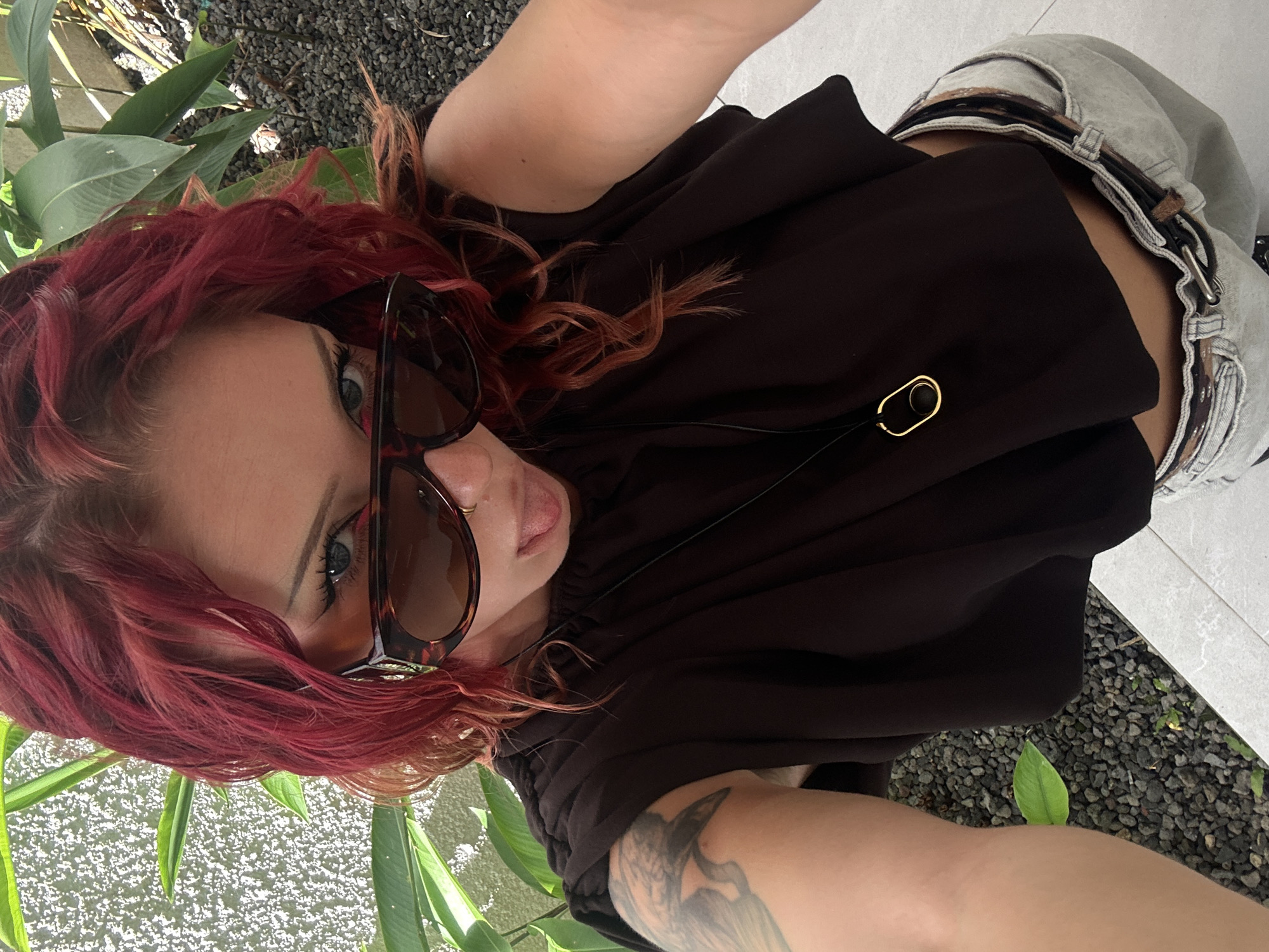 A woman with red hair and sunglasses is taking a selfie. She is wearing a dark brown top and light grey denim pants. Her arm has a tattoo of a bird. The background shows plants and grey gravel. The location is North Kuta, Indonesia.