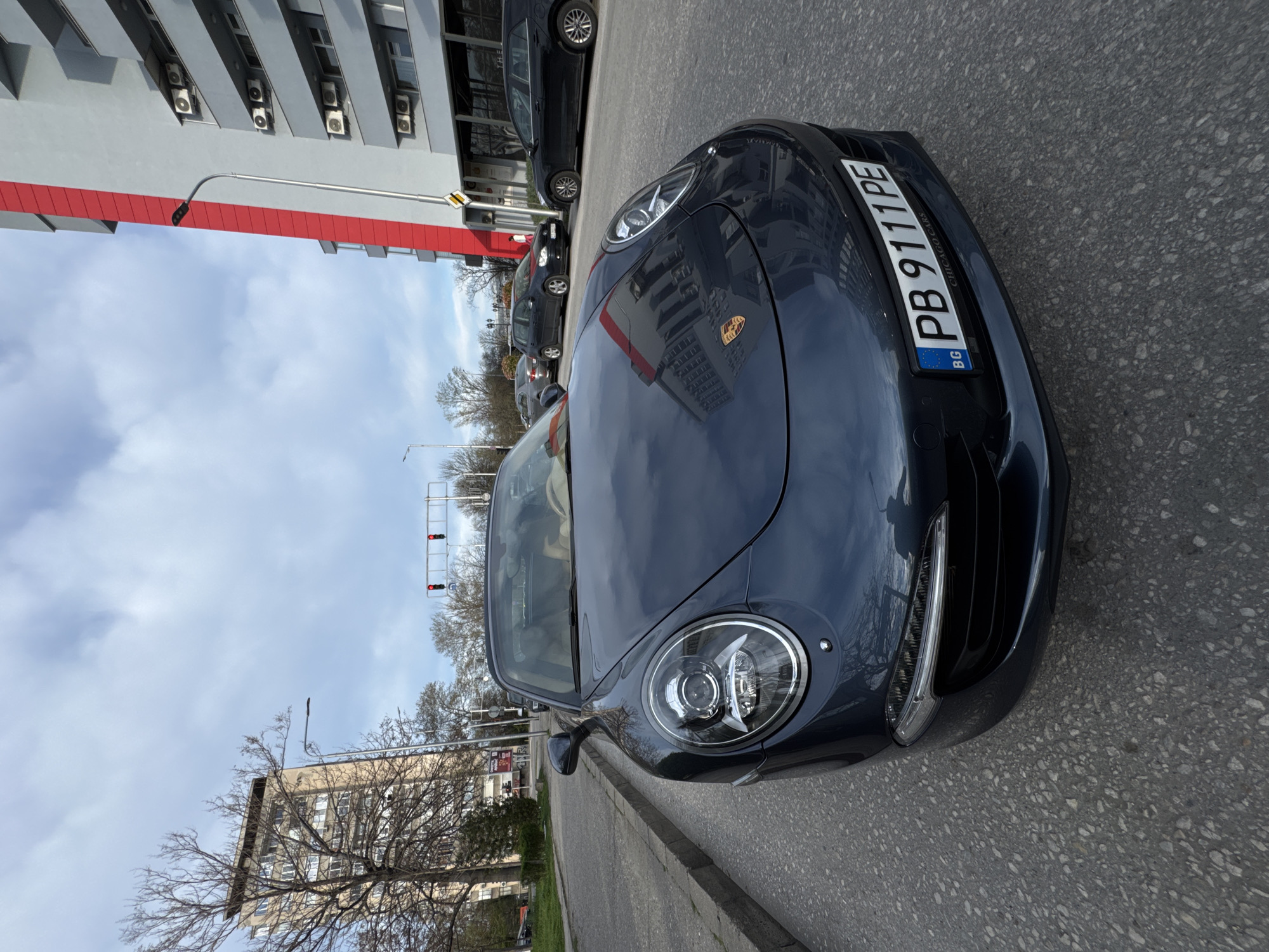 The image shows a gray Porsche 911 parked on the street in Plovdiv, Bulgaria. The car's license plate reads "PB 9111 PE". The background includes buildings, trees, and other parked cars. The sky is cloudy. A traffic light is visible in the background. The overall scene appears to be a typical urban streetscape.