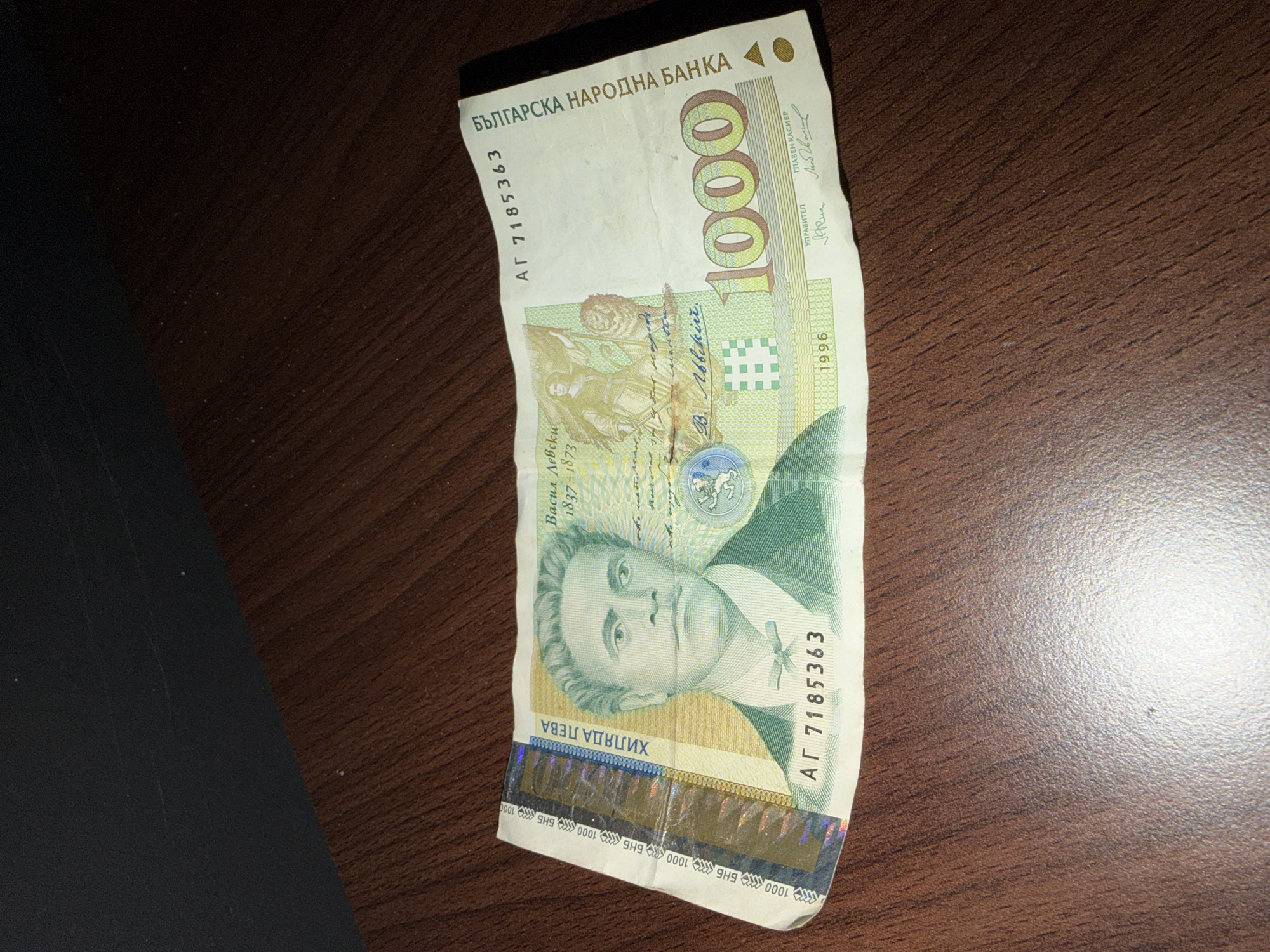 The image shows a single 1000 Bulgarian Lev banknote, dated 1996, lying on a wooden surface. The banknote features an image of Vasil Levski, a Bulgarian revolutionary, and the text "БЪЛГАРСКА НАРОДНА БАНКА" (Bulgarian National Bank). The serial number on the banknote is АГ 7185363. The bill is slightly crumpled and worn. There is no indication that the photograph was taken in Radomir, Bulgaria.