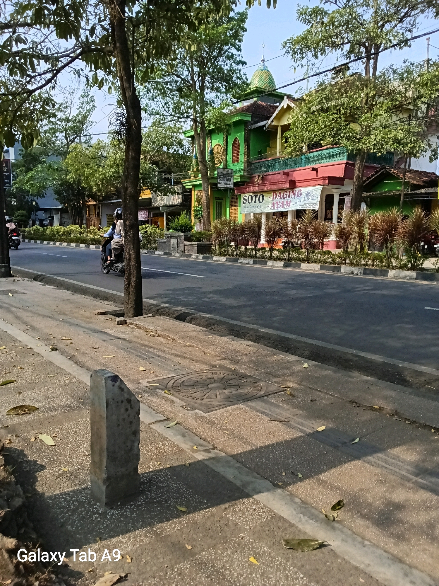 The image captures a street scene in the city of Kelutan, Indonesia. In the foreground, a concrete sidewalk is visible, with a stone marker and fallen leaves scattered across its surface. The road runs alongside the sidewalk, with a single lane of traffic. A motorcycle carrying two people is traveling on the road. On the opposite side of the road, there are buildings with vibrant green and red facades, and a green dome on one of the structures, suggesting it might be a place of worship. A banner advertising "SOTO DAGING AYAM" (chicken meat soup) is displayed prominently on one of the buildings. Trees with lush green foliage line the street, casting shadows on the road and sidewalk. The sky is clear and blue, indicating a sunny day. The overall impression is that of a typical urban street in Indonesia on a bright afternoon.