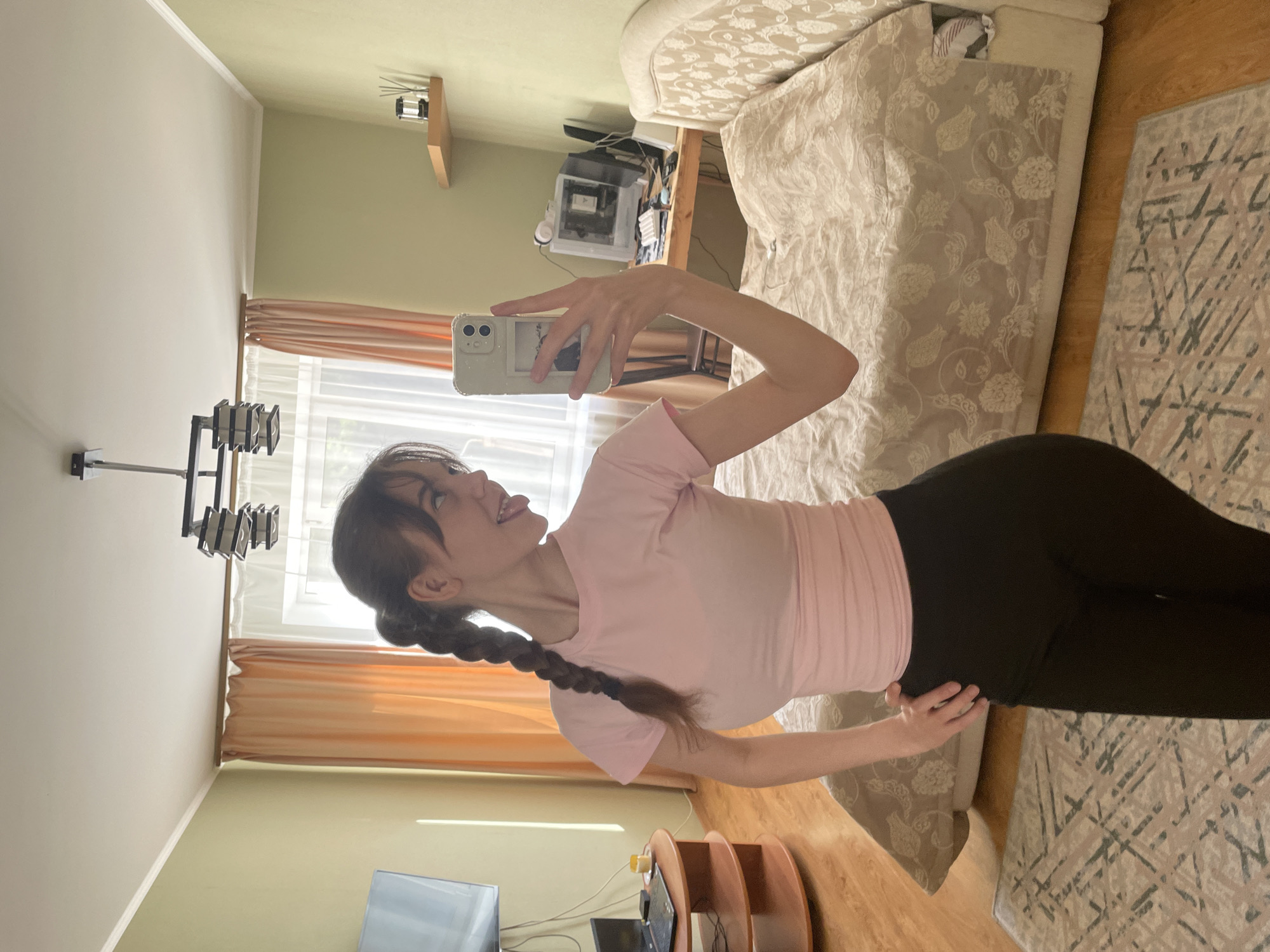 This image captures a young woman taking a mirror selfie indoors, likely in a bedroom or living space in Ufa, Russia. The photograph is rotated 90 degrees clockwise.
The subject is a young woman with dark hair styled in a long braid resting over her left shoulder. She is wearing a light pink, short-sleeved t-shirt and black leggings. She holds a white smartphone with a clear case, which has a small, indistinguishable picture or sticker on the back, in her right hand, angled to take the photo. Her left hand is placed on her lower back or hip. She exhibits a playful expression, with her tongue slightly sticking out.
The room is brightly lit by natural light streaming in from a window, indicating it is daytime. The window features sheer white curtains and peach-colored blinds or drapes. The walls are painted in two tones: a light beige on the left and a pale green on the back wall. The flooring is light-colored wood.
Various objects furnish the room: A large bed with an ornate, floral-patterned duvet cover and matching upholstered headboard is visible on the right. A patterned rug with geometric designs lies on the wooden floor beside the bed. In the background, a wooden shelf holds a computer tower, possibly a gaming PC, with visible internal components. Another small wooden shelf is mounted higher on the wall. On the far left wall, a modern black wall-mounted light fixture with multiple square lamps is installed. Towards the bottom left, a portion of a television screen and a small, circular, tiered wooden stand holding electronic devices and cables are visible.
No discernible text is visible on any objects in the image. The scene suggests a casual and personal moment captured in a well-lit interior setting.