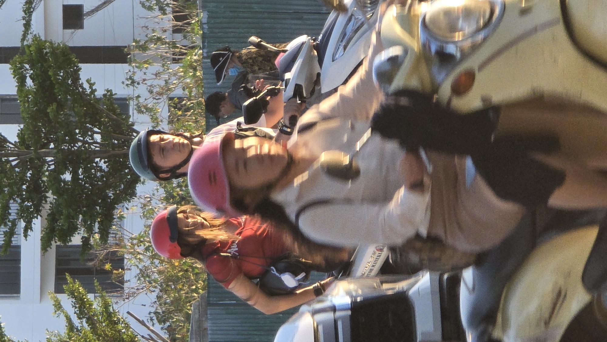 The image shows several people on motorbikes in Nha Trang Ward, Vietnam. A woman with a pink helmet is in the foreground, riding a motorbike. Behind her is another woman in a red shirt and a red helmet, also on a motorbike. Further back, a person with a green helmet can be seen. In the background, there is a building and trees, indicating an urban environment. Some people are wearing face masks. The overall scene suggests a busy street with people commuting on motorbikes.
