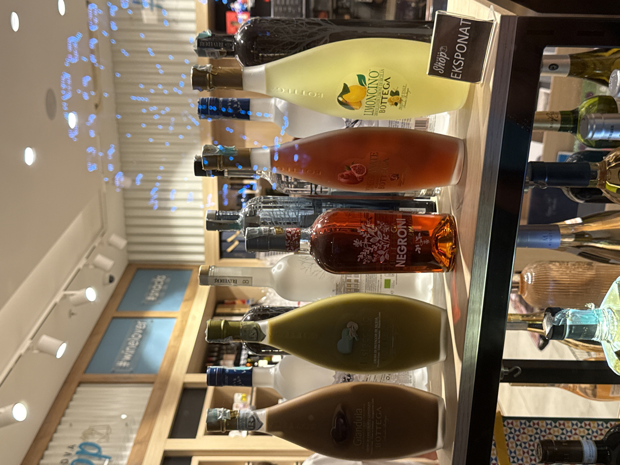 The image shows a retail display of alcoholic beverages inside a shop. Several bottles of liqueurs and spirits are arranged on shelves. In the foreground, on a wooden shelf, are several bottles with colorful liquids. From left to right, a chocolate liqueur labeled "Glanduia Bottega", a pistachio liqueur labeled "Il pistacchio", a dark red liqueur labeled "NEGRONI BOTTEGA", and a pale yellow liqueur labeled "LIMONCINO BOTTEGA". Behind these, on a higher shelf, are various clear and colored bottles, including what appears to be vodka. A small black sign with white text reading "Shop EKSPONAT" is placed on the right side of the foreground shelf. The background shows a wall with striped wallpaper and decorative blue lights. The overall environment suggests a well-lit store, possibly a duty-free shop or a specialty liquor store, given the prominent placement of the bottles and signage. Based on the presence of the sign "EKSPONAT" and the mention of Budva, Montenegro, this scene is likely located in Budva, Montenegro, possibly at a shop within a shopping center or a standalone store catering to tourists.