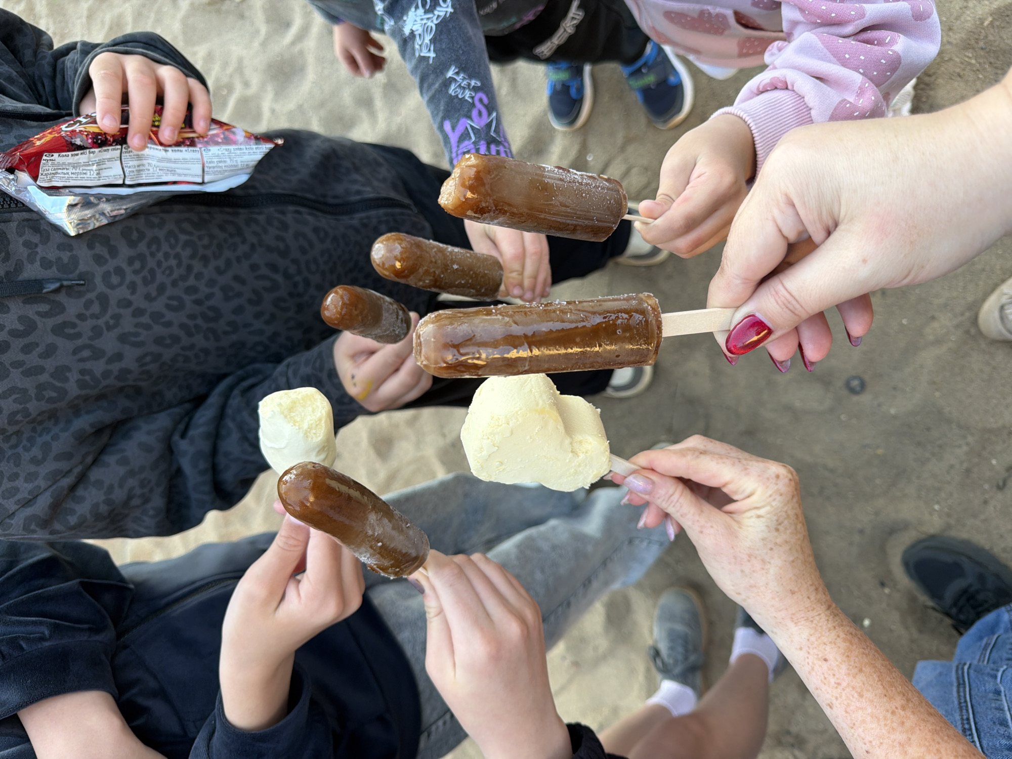This high-angle, top-down image captures a casual outdoor gathering of several individuals, including adults and children, sharing ice creams and popsicles. The scene is set on sandy or light dirt ground, suggesting a beach or park environment in Chaikovsky, Russia.
Six ice creams are prominently featured, held by various hands. Five are dark, translucent popsicles, possibly cola or caramel flavored, some exhibiting a frosty, textured surface. One is a creamy, light yellow ice cream bar, likely vanilla.
Visible clothing includes a dark grey/black animal print jacket, a pink long-sleeved top with white patterned dots, dark pants, and light blue jeans. Several pairs of sneakers (dark and grey) and white socks are also seen. One child holds a red and white snack bag.
Notable details include the presence of nail polish on adult hands: one with dark red/burgundy polish and another with a lighter, possibly iridescent or pastel shade. An arm with numerous freckles is also visible. Text on the snack bag, which appears to be in Kazakh and Russian, includes "«Кола хош иісі бар..." (Cola flavored...), "Таза салмағы: 70 г" (Net weight: 70 g), "Жарамдылық мерзімі 12 ай" (Shelf life: 12 months), and "coles", as well as "Проматен колы ассагу razy sterkt". A partial phrase "KEEP YOUR S" is visible on a child's sleeve. The overall activity conveys a sense of shared enjoyment, likely on a warm day.