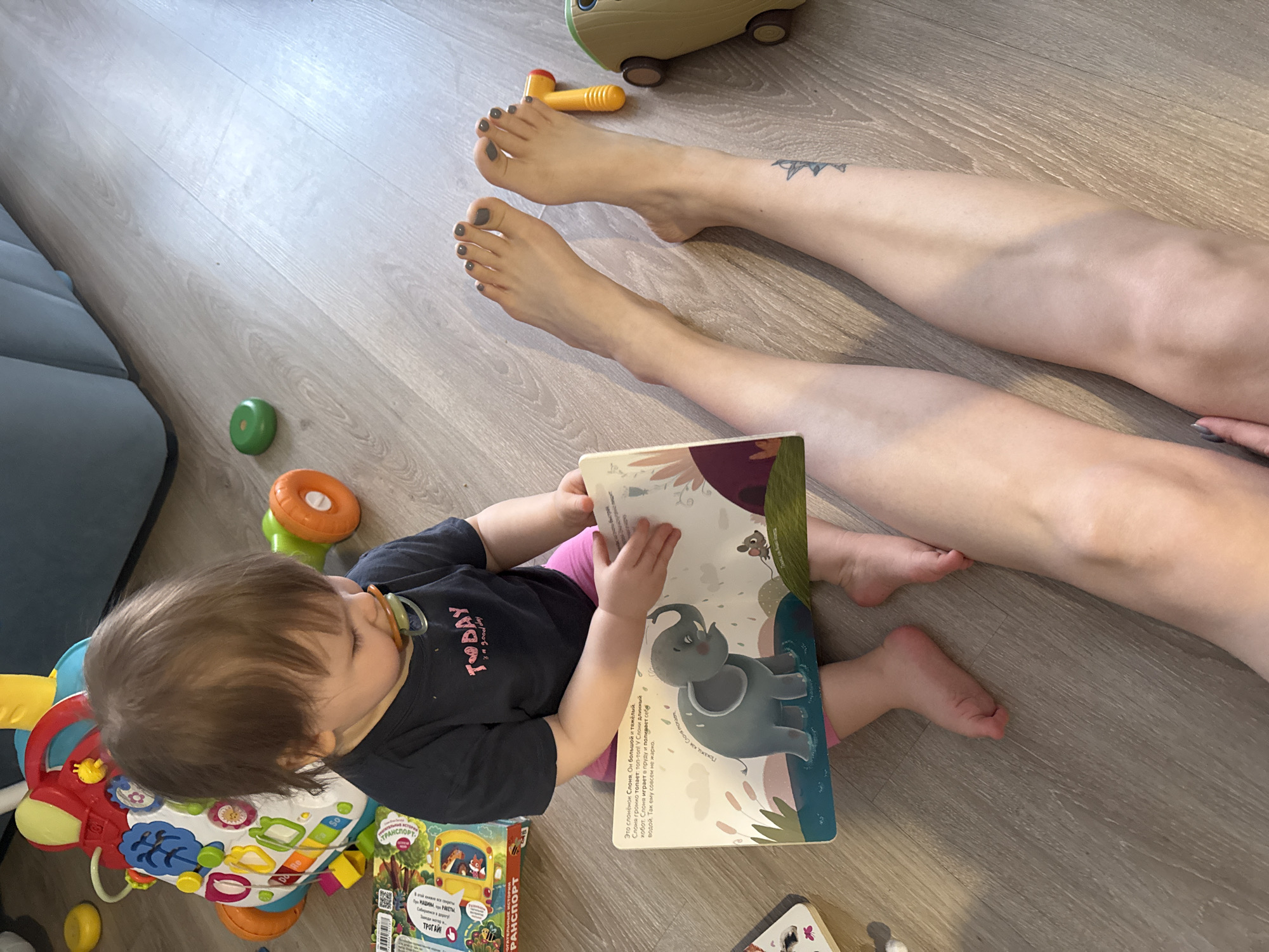 A baby lies on a wood-patterned floor with a blue sofa section on the left. The baby wears a navy shirt with a graphic on it, a pink bottom, and has a pacifier. They are surrounded by toys and a book with an elephant on the cover. A woman's legs are extended nearby, with gray-painted toenails and a tattoo on the ankle. The room is lit by ambient light. There is no explicit evidence of a Moscow, Russia, location.
