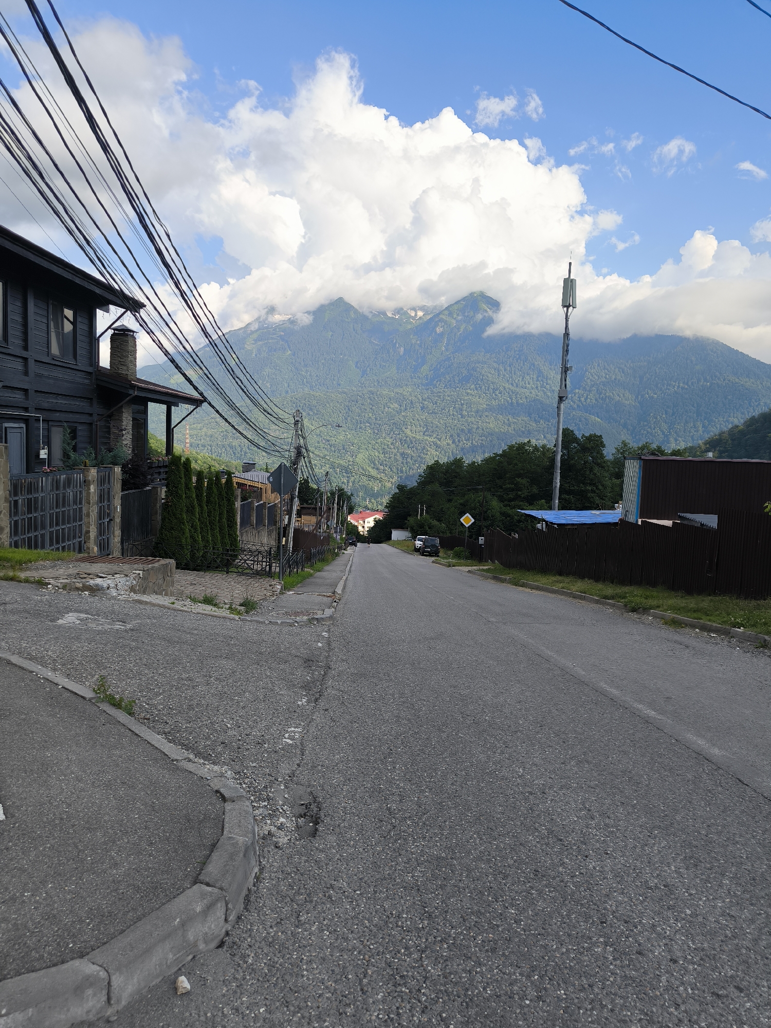 The image depicts a downward sloping asphalt road in Krasnaya Polyana, Russia, with a residential building on the left and a dense green mountain range in the background under a partly cloudy sky. The building on the left is dark wood with a stone chimney and a metal fence. Numerous overhead power lines crisscross the scene, supported by utility poles. A cellular tower is visible on the right side of the road, positioned in front of the mountains. The road surface shows signs of wear, with cracks and gravel. Several trees and fences line the sides of the road, and in the distance, a few other houses can be seen. The overall environment suggests a mountainous, somewhat rural area within a settlement.
