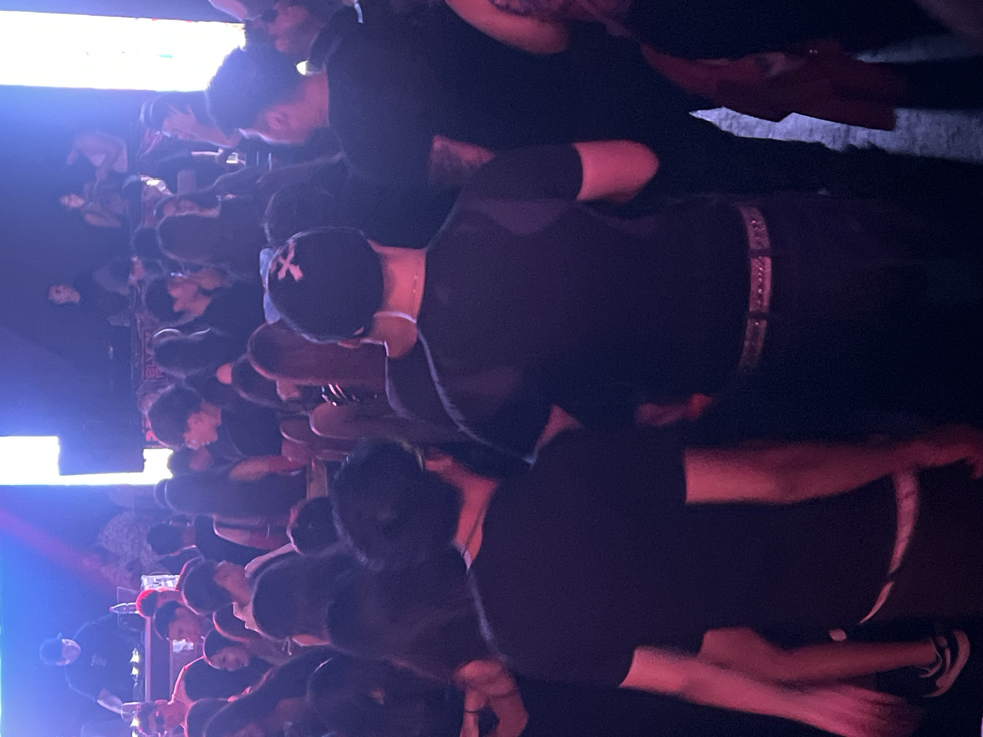 The image shows a crowded indoor space, likely a concert venue or nightclub, in Arica, Chile. The scene is dimly lit with blue and red lighting. Many people are visible, mostly in the center of the image, facing the same direction, presumably toward a stage. Many are wearing dark clothing, possibly black t-shirts. A few people are wearing caps. The crowd appears dense, with people close together. The floor is not visible.