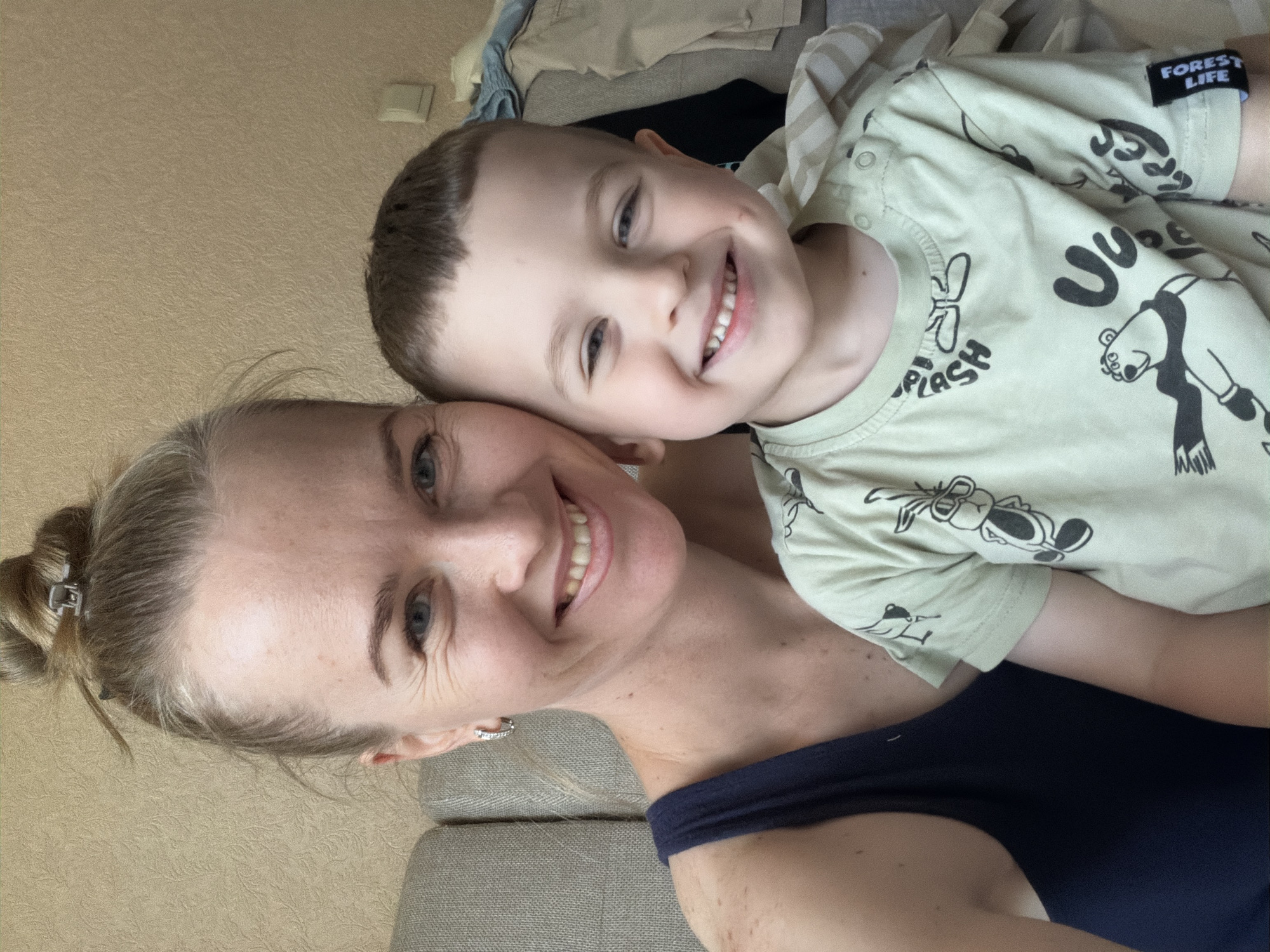 A woman with blonde hair pulled into a bun and a young boy with short brown hair are posing for a selfie in what appears to be a living room in Khabarovsk, Russia. The woman is wearing a navy blue tank top and a sparkling earring. The boy is wearing a light green t-shirt with cartoon characters on it, including a bear and what appears to be a rabbit. Both are smiling and looking at the camera. They are seated on a couch, with a textured wall behind them. Clothing is visible on a chair or sofa in the background.