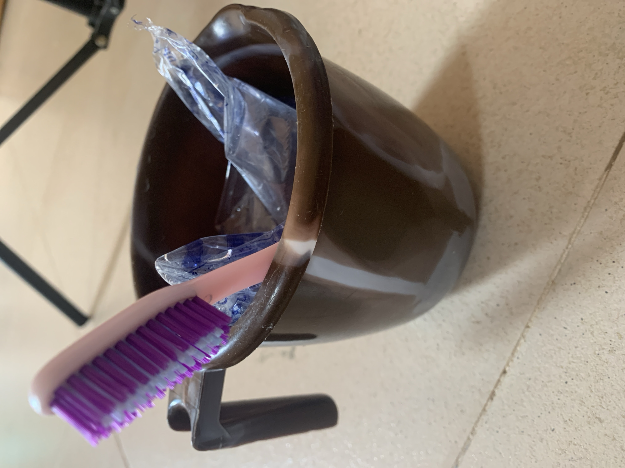 A toothbrush with a pink handle and purple bristles rests inside a brown mug. The mug also contains two plastic-wrapped items with blue contents. The mug is situated on a tiled floor. A black tripod leg is visible in the background.