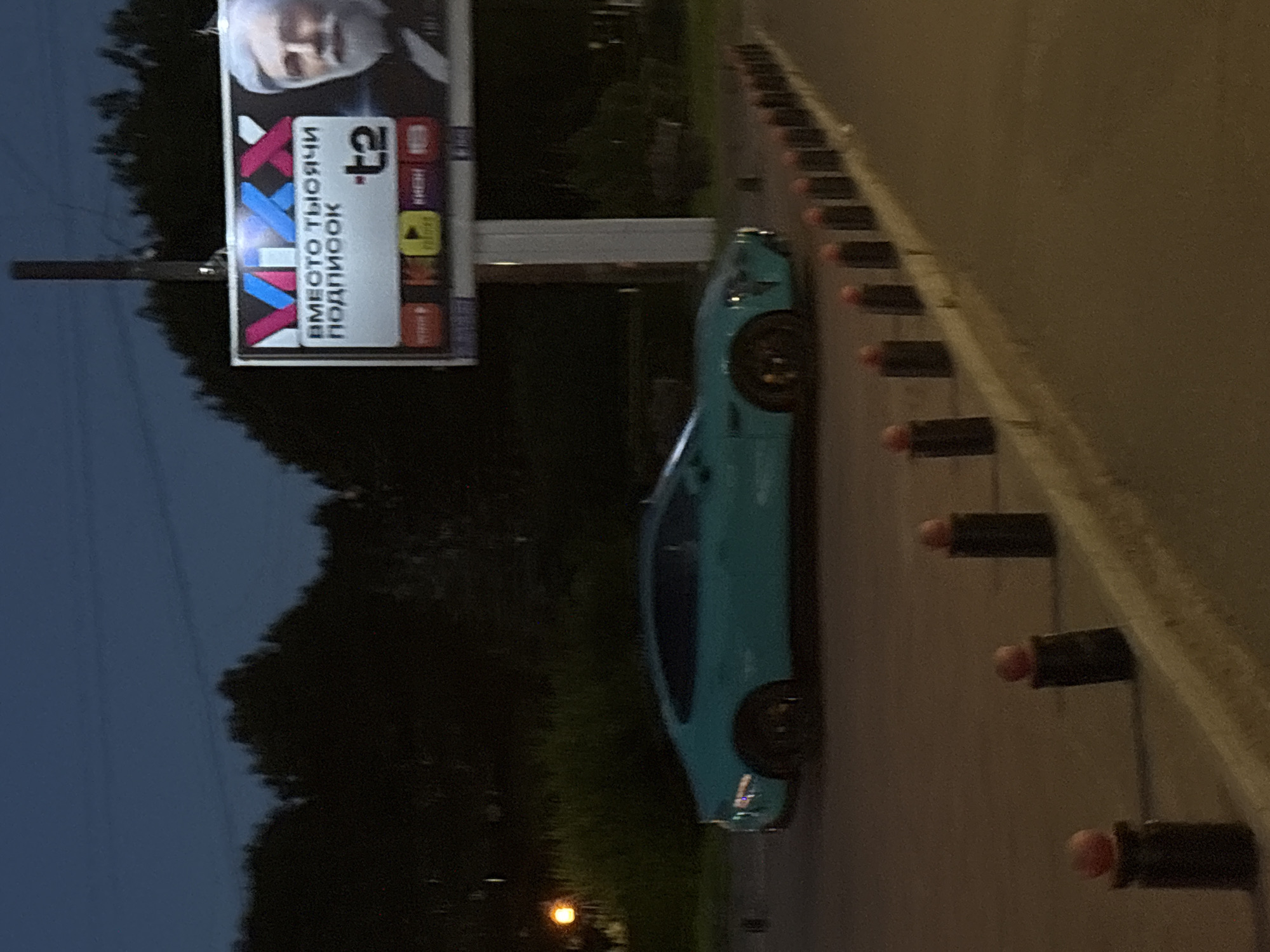 A turquoise-colored sports car is parked on the side of a road at dusk. The car has black wheels and is angled towards the viewer. In the background, a billboard is visible with text in Russian and images, suggesting an advertisement. The billboard features the letters "VXX" in large, colorful font, with the text "ВМЕСТО ТЫСЯЧИ ПОДПИСОК" below it, which translates to "Instead of a thousand subscriptions." To the right of this text is the logo "t2". The scene is set against a dark, twilight sky and a silhouette of trees. A row of black bollards with red tops lines the edge of the road in the foreground, indicating a pedestrian walkway or separation. The lighting suggests it is an urban or suburban environment in Saint Petersburg, Russia.