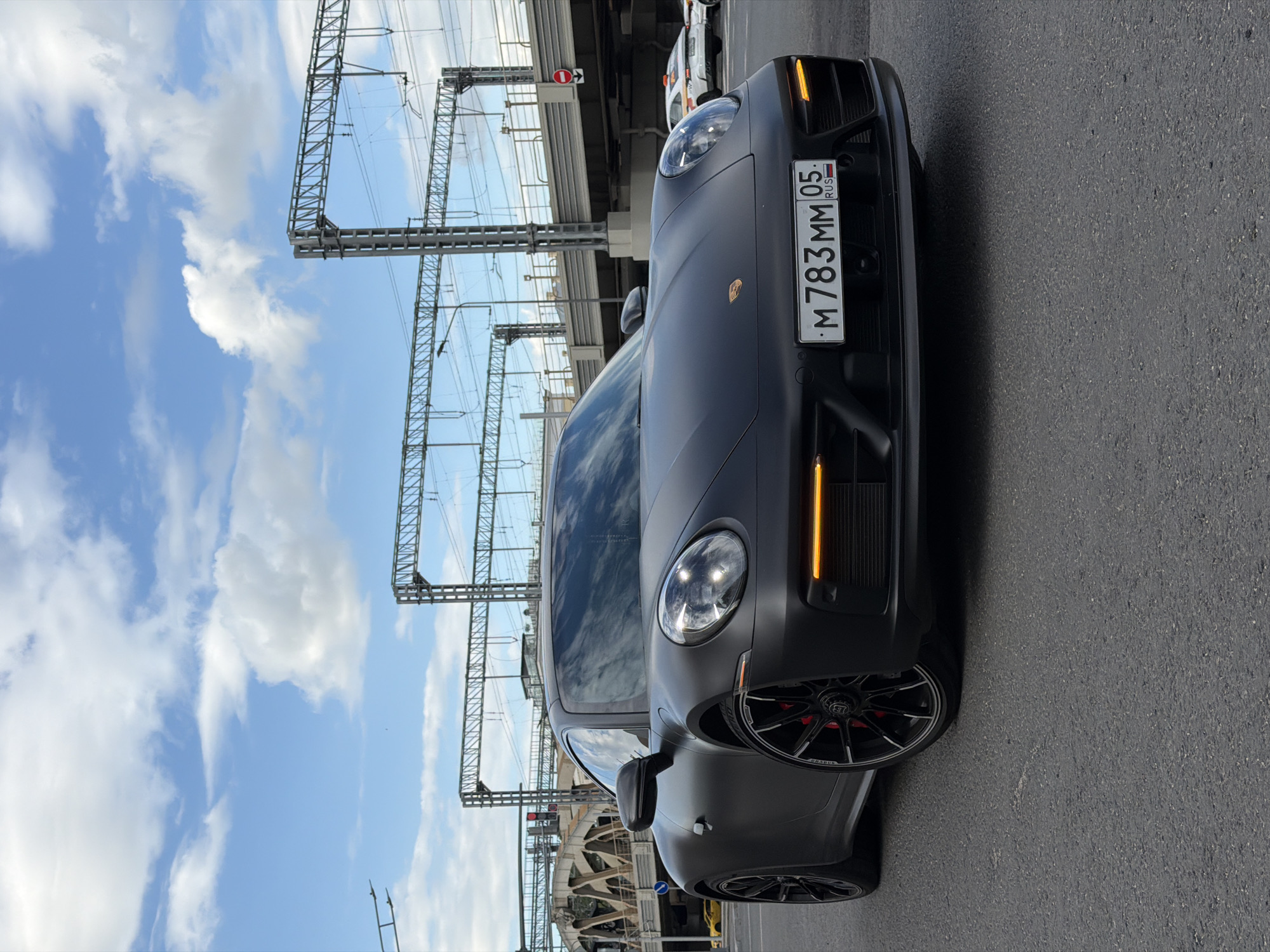 The image shows a matte gray Porsche Panamera parked on asphalt under a bridge structure with overhead power lines. The license plate reads "M 783 MM 05 RUS". A taxi can be seen further down the road under the bridge. The setting appears to be urban, possibly in Moscow, Russia. The sky is blue with some clouds.