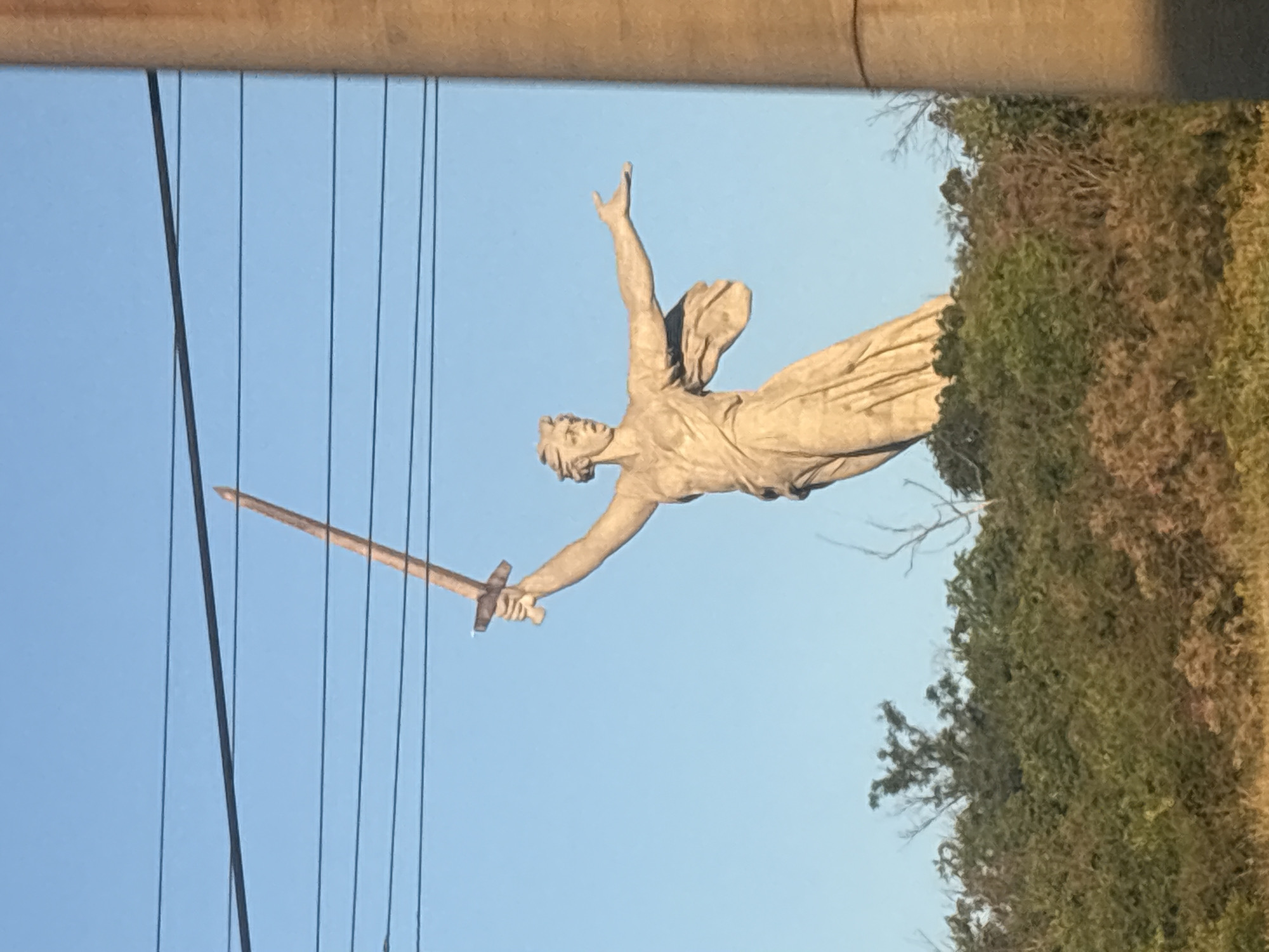 A monumental statue, "The Motherland Calls," stands tall against a clear blue sky in Volgograd, Russia. The statue depicts a woman with her arms outstretched, one holding a large sword aloft. She is positioned atop a grassy hill covered with dense foliage. Several power lines are visible in the upper portion of the frame, crossing horizontally against the sky. To the right, a portion of a large cylindrical concrete pillar is present. The statue is made of concrete and appears to be weathered.