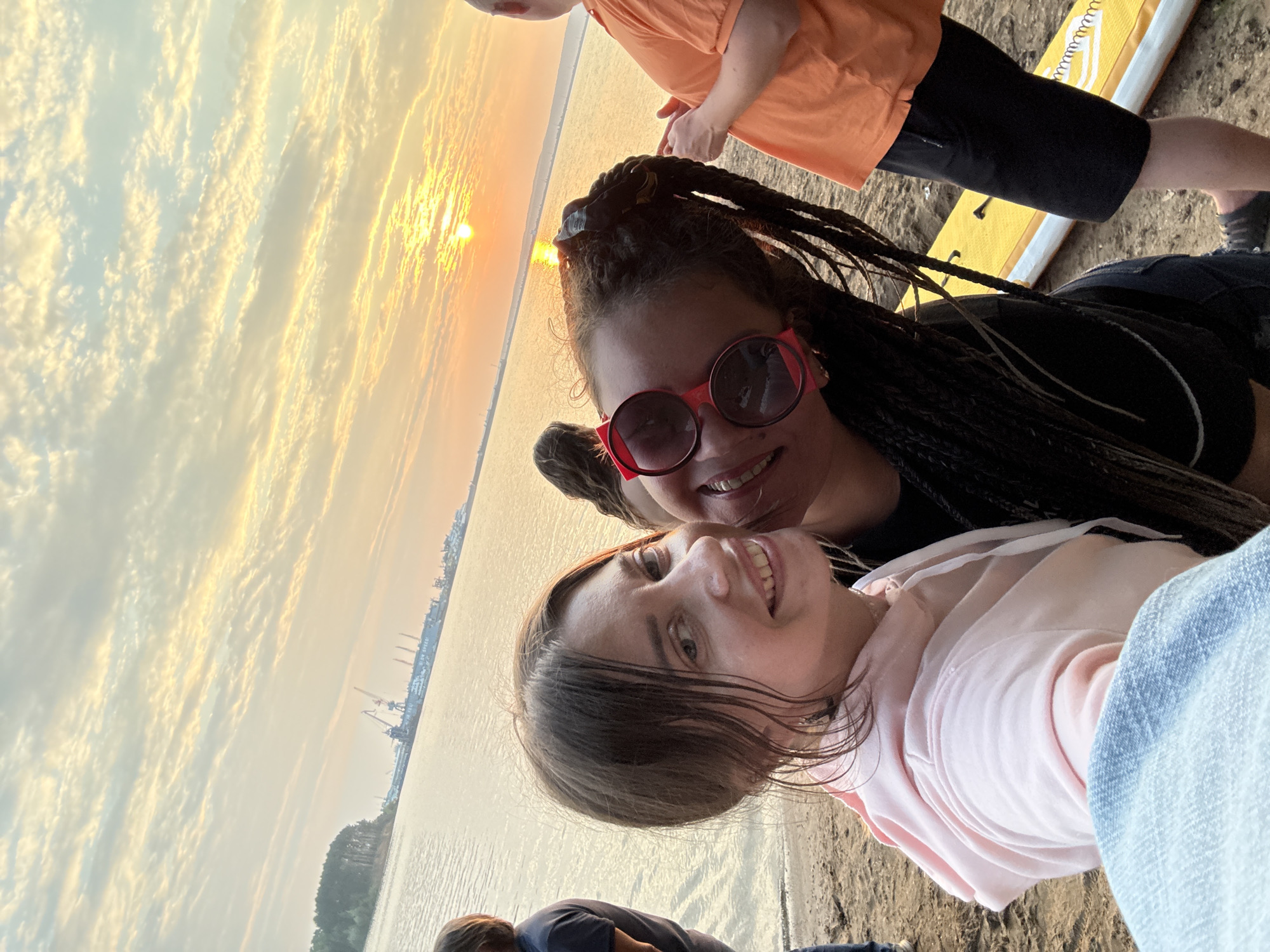 Two women are taking a selfie on a sandy beach at sunset. The woman in the foreground on the left is smiling at the camera, wearing a pink hoodie and blue jeans. The woman on the right is also smiling, wearing large red sunglasses and her hair is in braids. Behind them, the sun is setting over a body of water, casting a warm glow across the sky. In the distance, there is a shoreline with trees and a port with cranes. Another person in an orange shirt is visible on the right side of the frame, standing near a yellow object that appears to be a paddleboard. The overall scene suggests a relaxed, outdoor recreational activity.