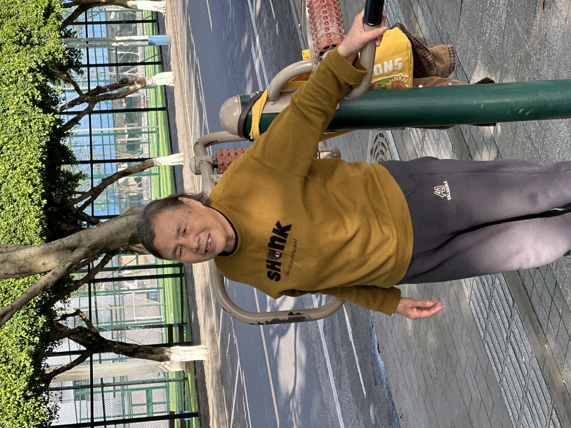 An elderly individual, likely a woman, is engaged in light exercise at an outdoor public park in Jiangjin, China. She is positioned next to a piece of green and grey outdoor fitness equipment, gripping a black handle with her left hand. Her right arm hangs by her side, and she is looking towards the camera with a gentle smile.
She is dressed in a mustard yellow long-sleeved sweater that features black text reading "SHONK" with a small circular emblem, and beneath it, "use dior by the mind". Her lower body is clad in dark grey sweatpants with white text that reads "A1 VALUABLE". A yellow bag, possibly with a sunflower pattern, is hanging on the exercise equipment near her left arm, and a brown garment is tucked behind it. The equipment itself includes textured red rollers, often used for massage or flexibility.
The setting appears to be a bright, clear day, indicated by the well-lit scene and visible shadows. In the background to her left, lush green foliage and mature trees with white-painted trunks are visible. Beyond the trees, a tall green wire fence, possibly enclosing a sports court (like a tennis or basketball court), can be seen. To her right and extending into the background, there is a paved area with white demarcation lines, suggesting a path or another court. The ground in the immediate foreground where she stands is paved with grey interlocking tiles.