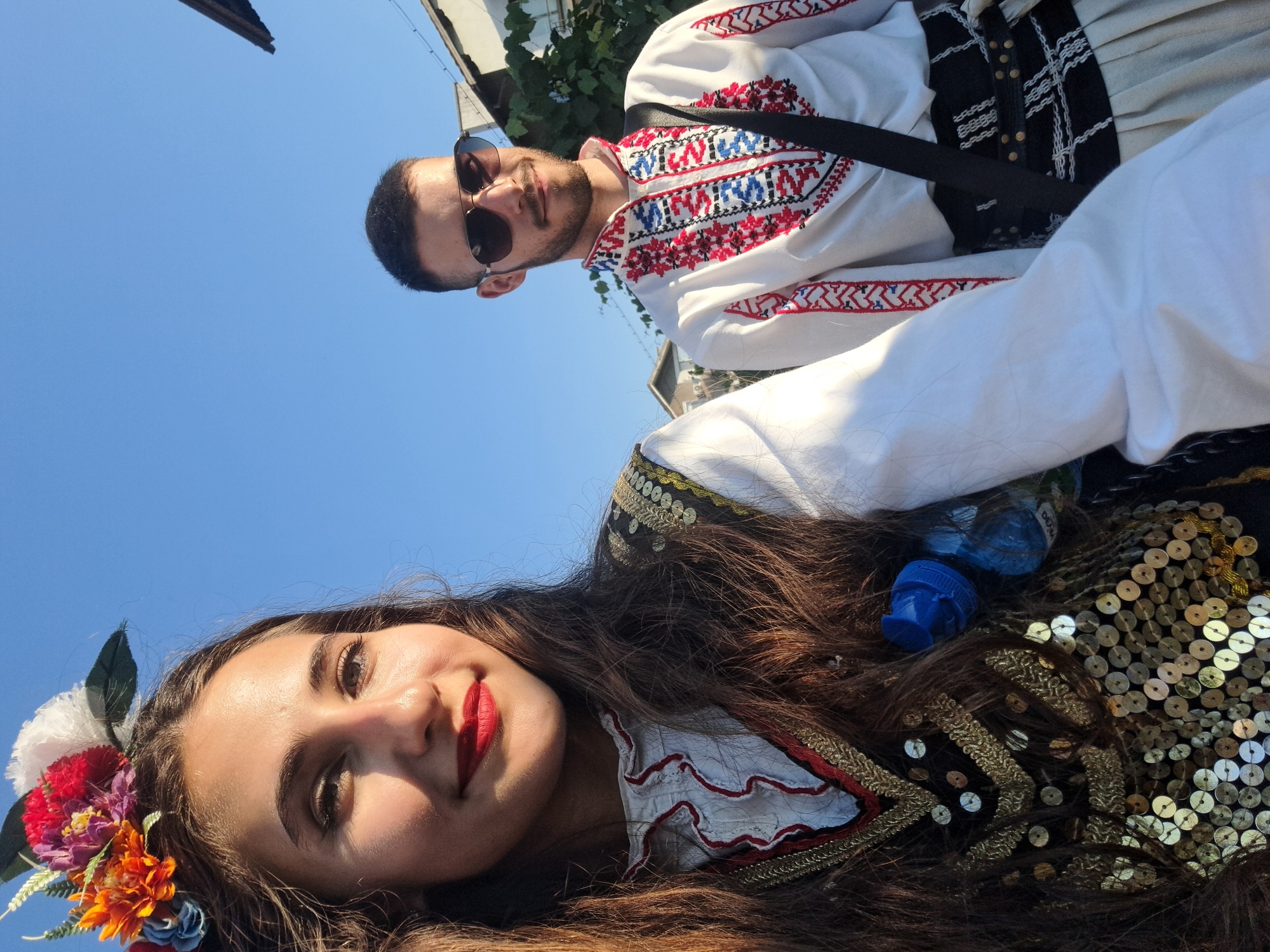 In an outdoor, daylight scene with a clear blue sky, a young woman with long, dark hair adorned with a floral headdress is pictured in the foreground, taking a selfie. She is wearing a white blouse with red and gold trim and a black vest embellished with gold sequins and trim. A blue water bottle is clipped to her clothing. To her right and slightly behind her, a young man is also visible. He has short, dark hair and is wearing aviator sunglasses and a white shirt with intricate red, blue, and black embroidery around the collar and down the front. He has a black strap across his chest and is wearing a wide, black belt with a decorative buckle over a light-colored pleated skirt. The background shows glimpses of buildings and greenery.