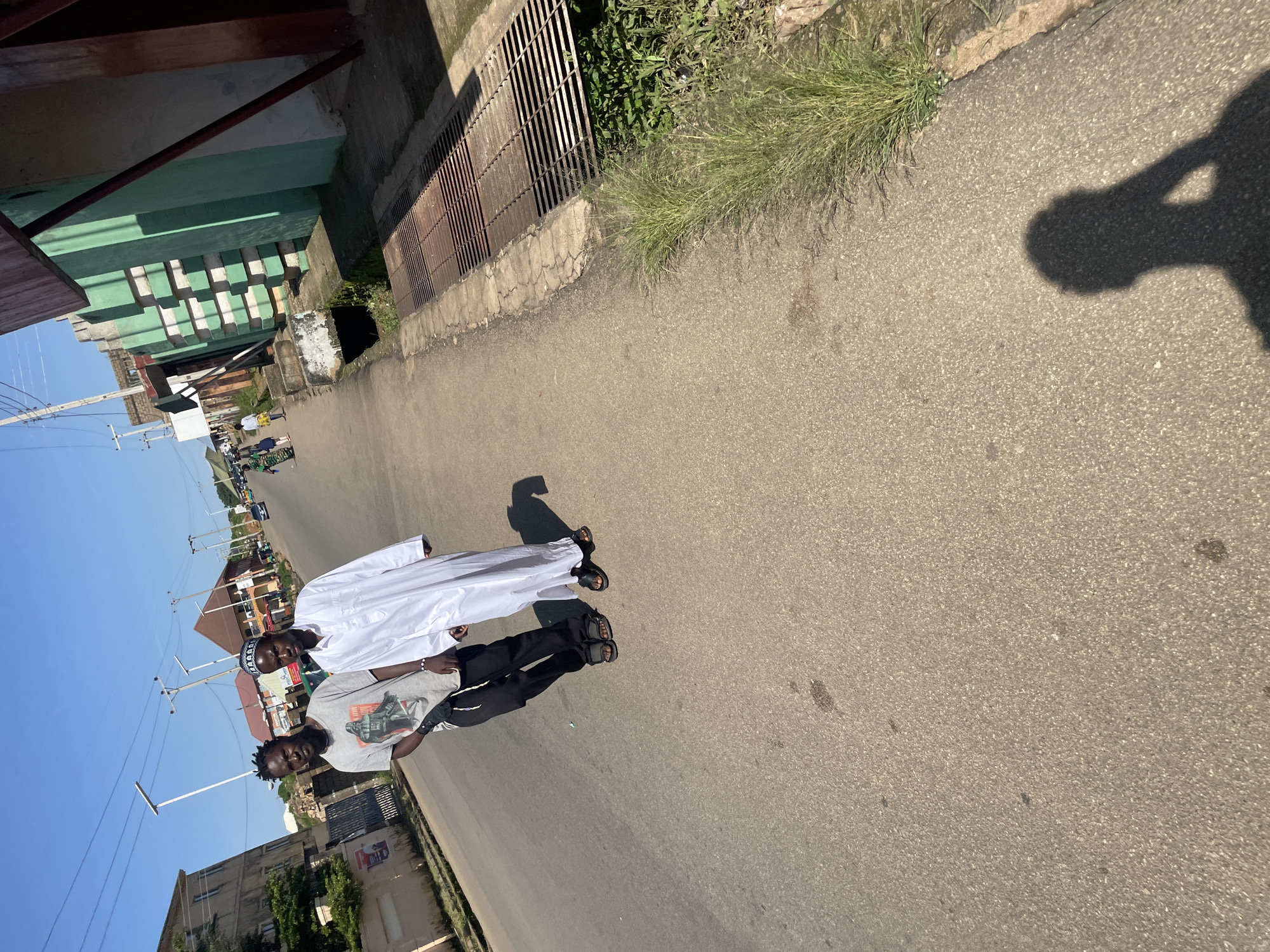 Two young men are walking on a paved road in Jos, Nigeria, on a clear, sunny day. The man on the left is wearing a grey t-shirt with a Darth Vader graphic and black pants. He has dark, braided hair and appears to be wearing dark sandals. The man on the right is wearing a white thobe and a kufi cap, suggesting he is Muslim. He is also wearing dark sandals. They are walking in the same direction, side-by-side, with the man on the right slightly ahead. The road is asphalt and appears to be a typical urban street in Jos, with buildings and power lines visible in the background. To the right of the road, there is a drainage grate and some overgrown grass. The shadow of the person taking the photo is visible in the foreground.