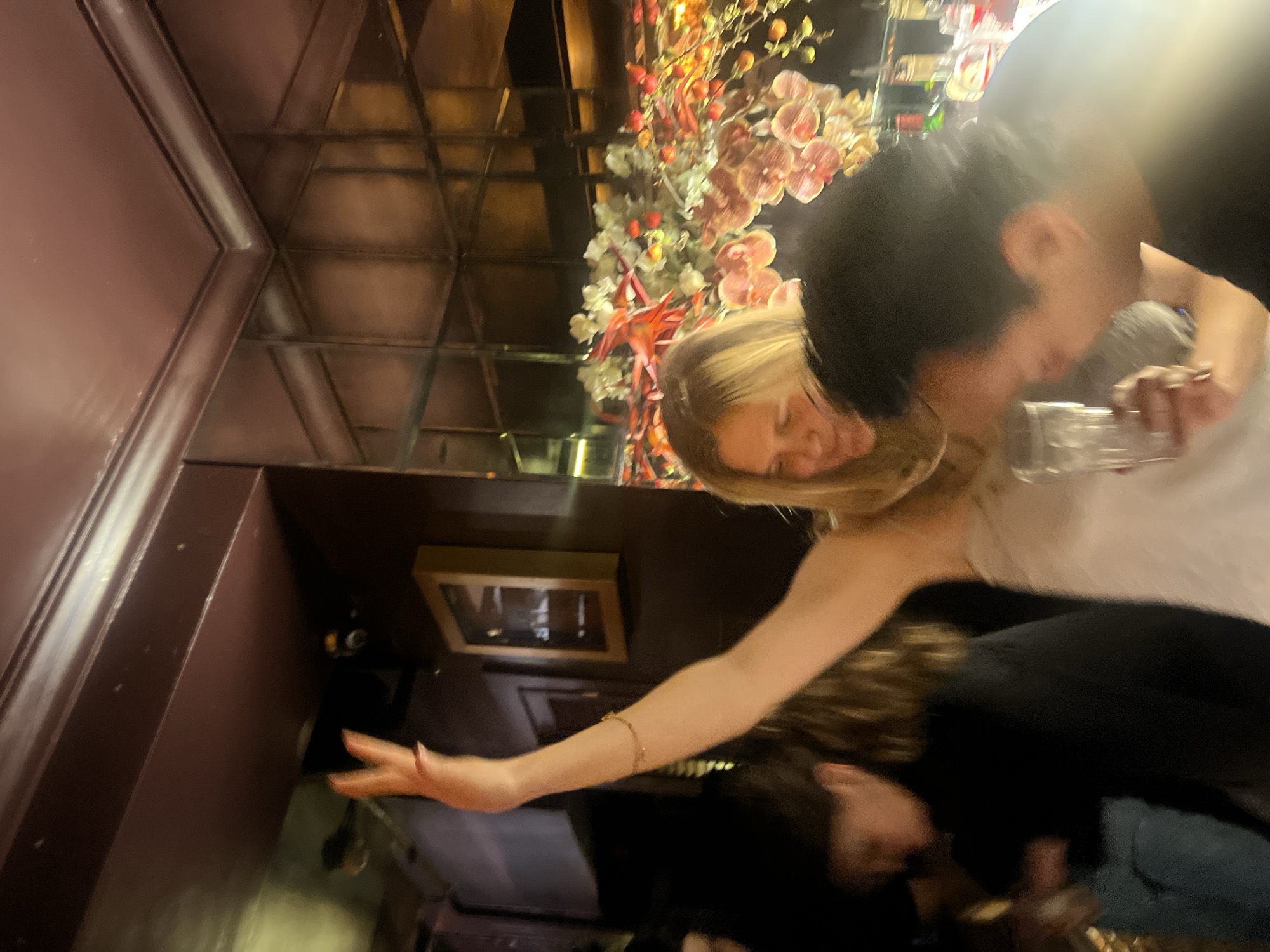 The image depicts an indoor scene, possibly a bar or restaurant in London, UK. A fair-skinned woman with blonde hair, wearing a white top and a gold bracelet, extends her arm, holding a glass. A dark-haired man leans close to her, also holding a glass. There is an ornate floral arrangement reflected in a mirrored ceiling. The environment includes dark paneled walls and what appears to be a framed picture. The lighting is dim.