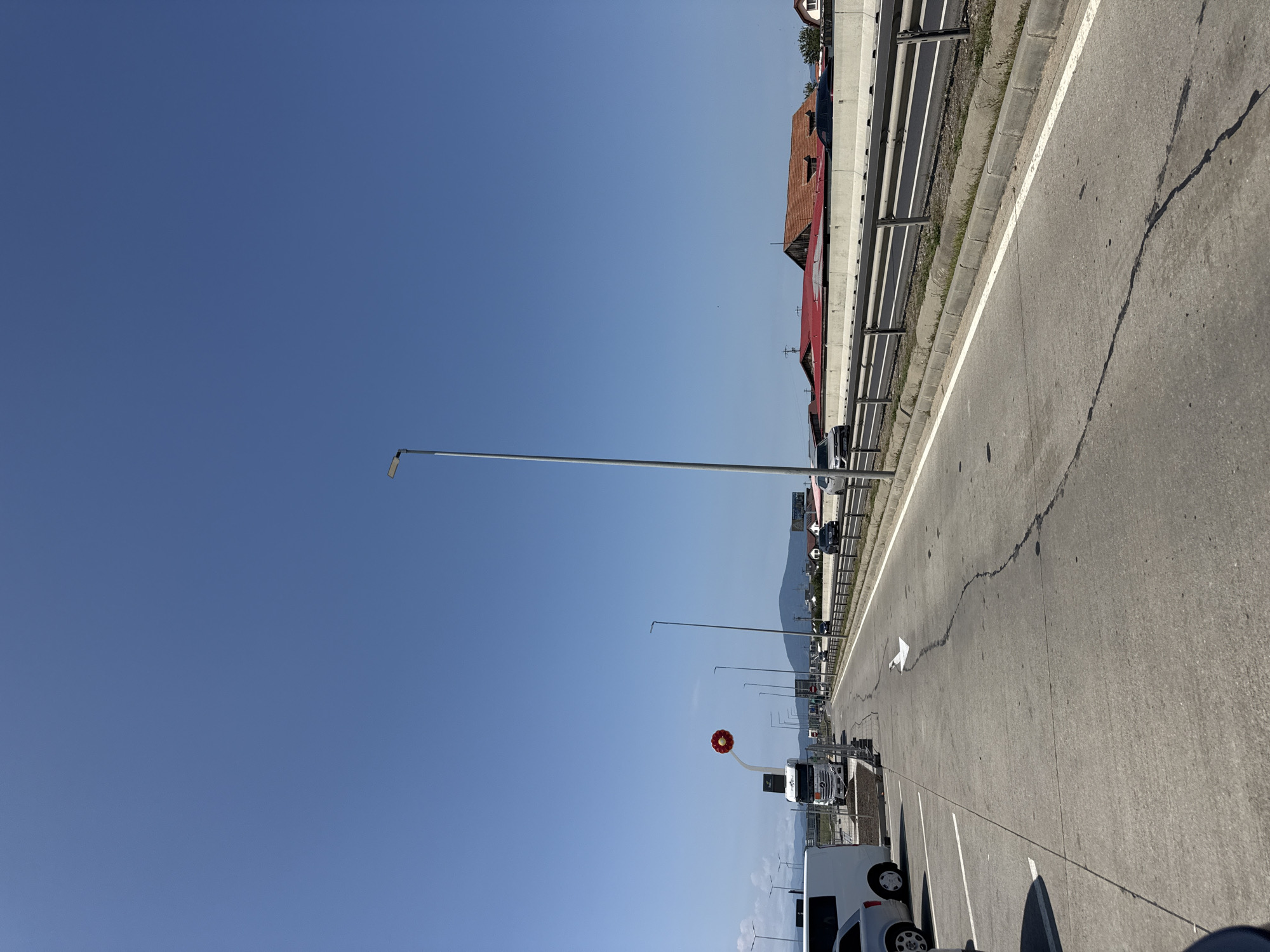 The image shows a view of a highway in Sanpetru, Romania, on a clear, sunny day. The highway has multiple lanes and is bordered by a concrete barrier with a metal guardrail. Several vehicles, including cars and a white truck, are visible on the highway. On the left side of the highway, there is a parking area with a white van and a silver car. A tall lamppost stands on the right side of the highway. In the background, there are buildings with red roofs and a hill with some vegetation. A large red and yellow circular sculpture is prominently displayed on a tall pole near the white truck. The ground of the parking area is made of concrete and shows some cracks. The sky is a clear, bright blue.