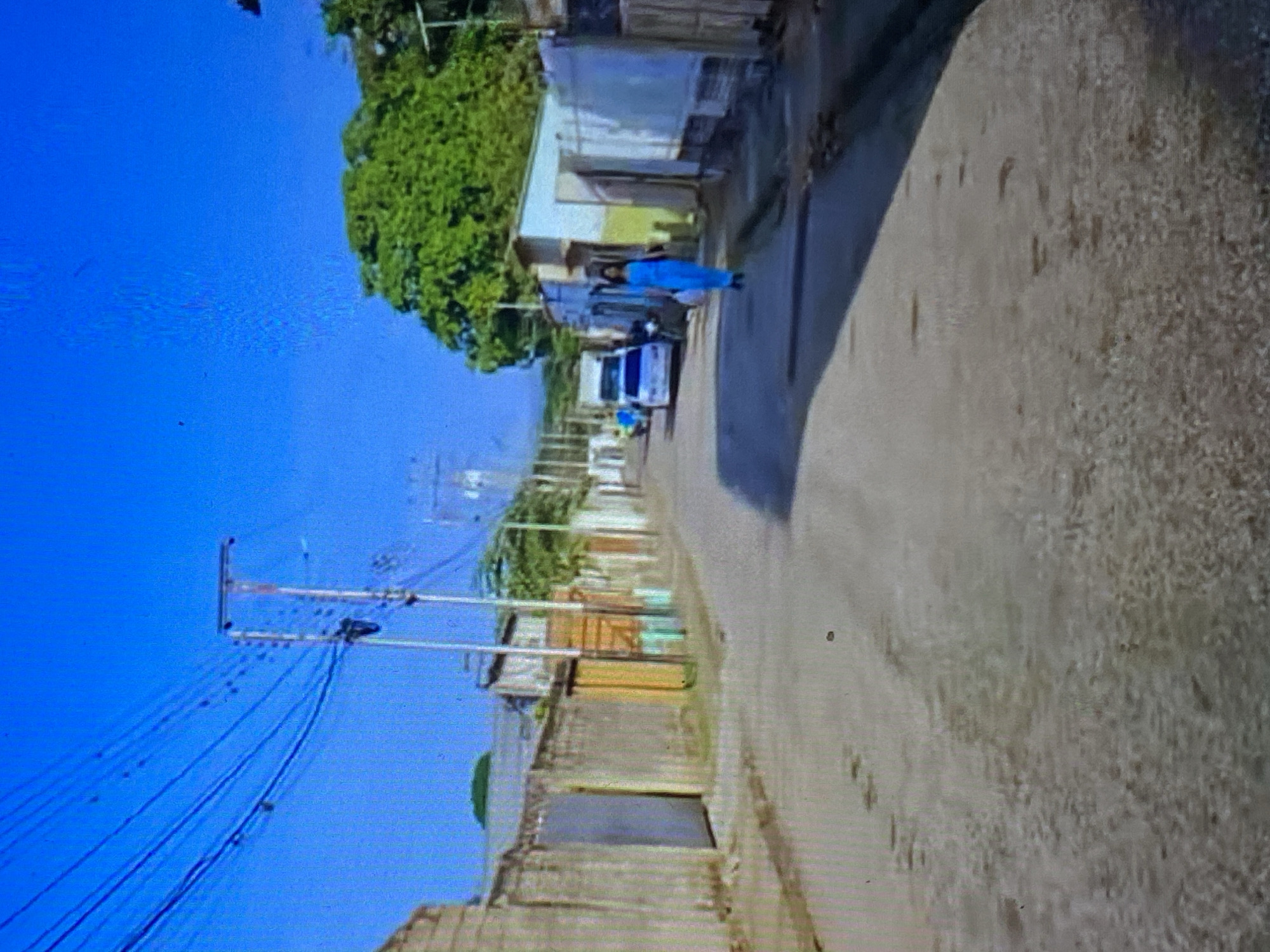 The image captures a bright, sunlit street scene in a residential or semi-urban area. The perspective is from the street, looking down a paved road that recedes into the distance.
In the mid-ground, a person wearing a striking bright blue outfit, possibly a uniform or scrubs, walks away from the viewer down the center of the road. Following or parked near them is a white van with blue accents, which could be a police, utility, or public service vehicle.
On the left side of the street, a series of simple, low-rise buildings line the road. These structures exhibit varying architectural styles, with some appearing to be constructed from concrete or stucco, showing different textures and subtle color variations. A large, verdant green tree stands prominently on the far left, its foliage contrasting with the clear blue sky.
The sky is a vivid, cloudless blue, indicating a bright daytime setting with sunny weather. Overhead on the left, a prominent network of utility poles and numerous power or telephone lines spans across the frame. The paved road surface appears to be asphalt or concrete, showing some wear and texture.
The scene conveys a sense of everyday life in a community, with the buildings and utility infrastructure suggesting a developed but perhaps modest urban environment. No legible text is visible on any objects or structures. The image quality suggests it might be a photograph taken of a screen, exhibiting some minor pixelation and display artifacts.