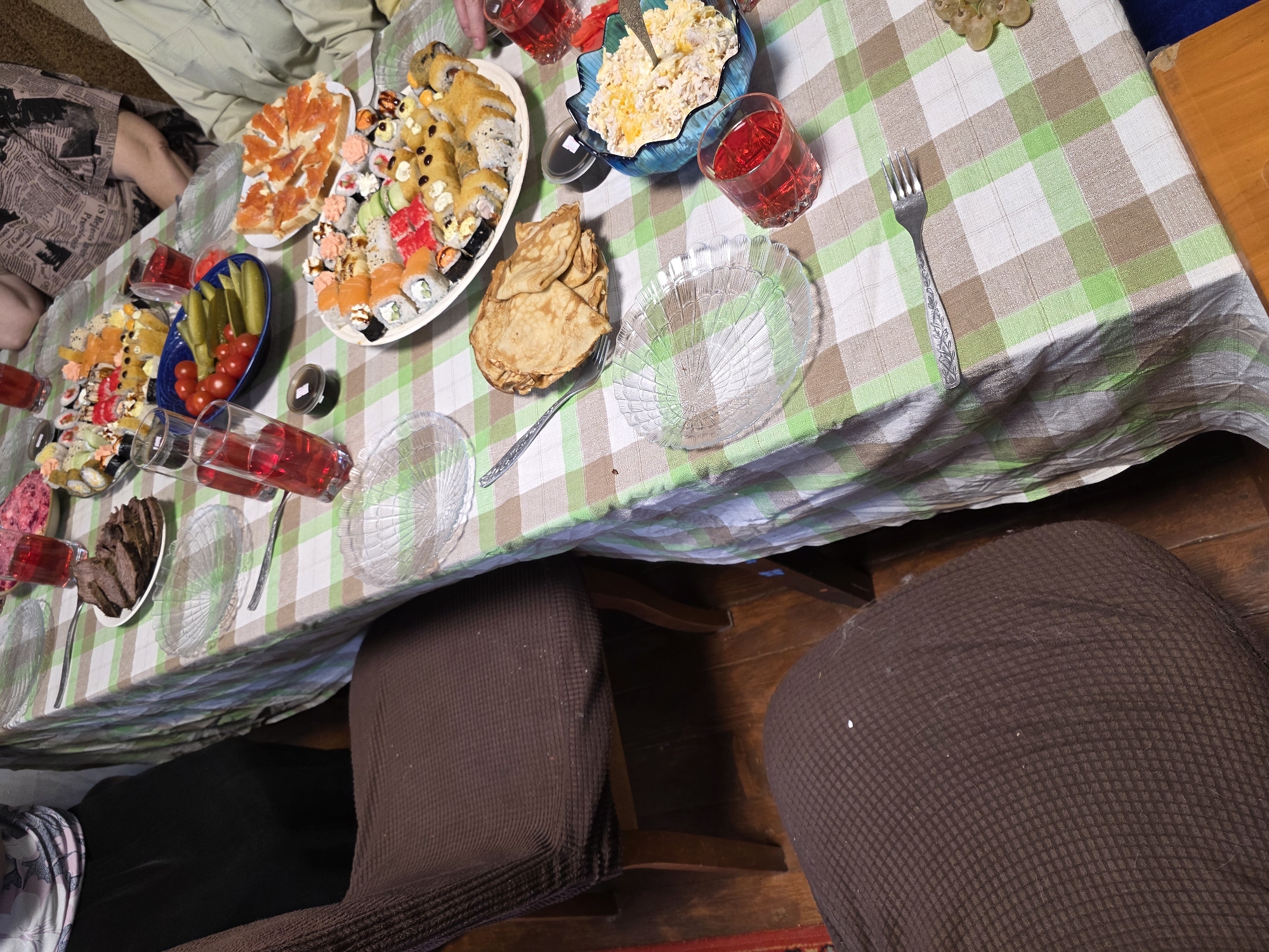 This image captures an indoor scene of a dining table set for a meal or gathering, likely in Chaikovsky, Russia. The table is adorned with a green, brown, and white checkered tablecloth. Several individuals are seated around the table, though only their torsos, arms, and legs are visible from this high-angle perspective.
The table is laden with a diverse spread of food. Prominently featured are two large platters of sushi and maki rolls, displaying various types, colors, and toppings. One platter also includes small bread appetizers topped with salmon. A blue bowl holds a creamy salad, and another blue dish contains a vibrant assortment of pickled cucumbers and fresh cherry tomatoes. A stack of golden pancakes or blini is present on a plate, along with a separate dish of sliced dark bread.
Clear glass plates, some with an ornate shell-like pattern, are distributed around the table, accompanied by several forks with decorative handles. Multiple clear glasses filled with a red beverage, possibly juice or compote, are also visible.
Below the table, the legs of the seated individuals and dark brown, textured chair covers can be seen. The flooring appears to be wooden. One person's attire includes a fabric with a newspaper print pattern, though no specific text is legible. The scene suggests a convivial atmosphere, indicative of a family meal or a social occasion, with a plentiful and varied selection of dishes. The time of day cannot be determined, and no specific emotions are discernible from the visible parts of the people.