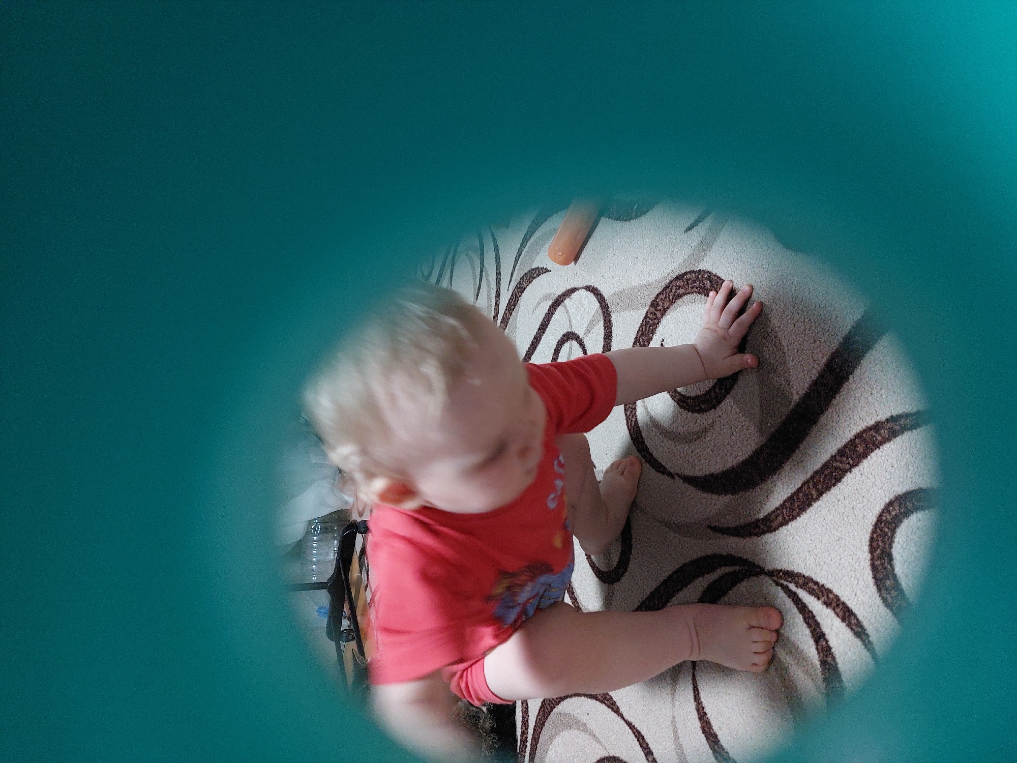 A view through a circular teal-colored opening reveals a toddler on a beige carpet with brown swirl patterns. The child, wearing a red t-shirt with a graphic, is positioned with their feet and right hand touching the carpet. The toddler's head is oriented towards the right of the frame. A table with items is visible in the background on the left, partially obscured by the teal frame. An orange object is visible on the carpet above the child. This photo was taken in Chaikovsky, Russia.