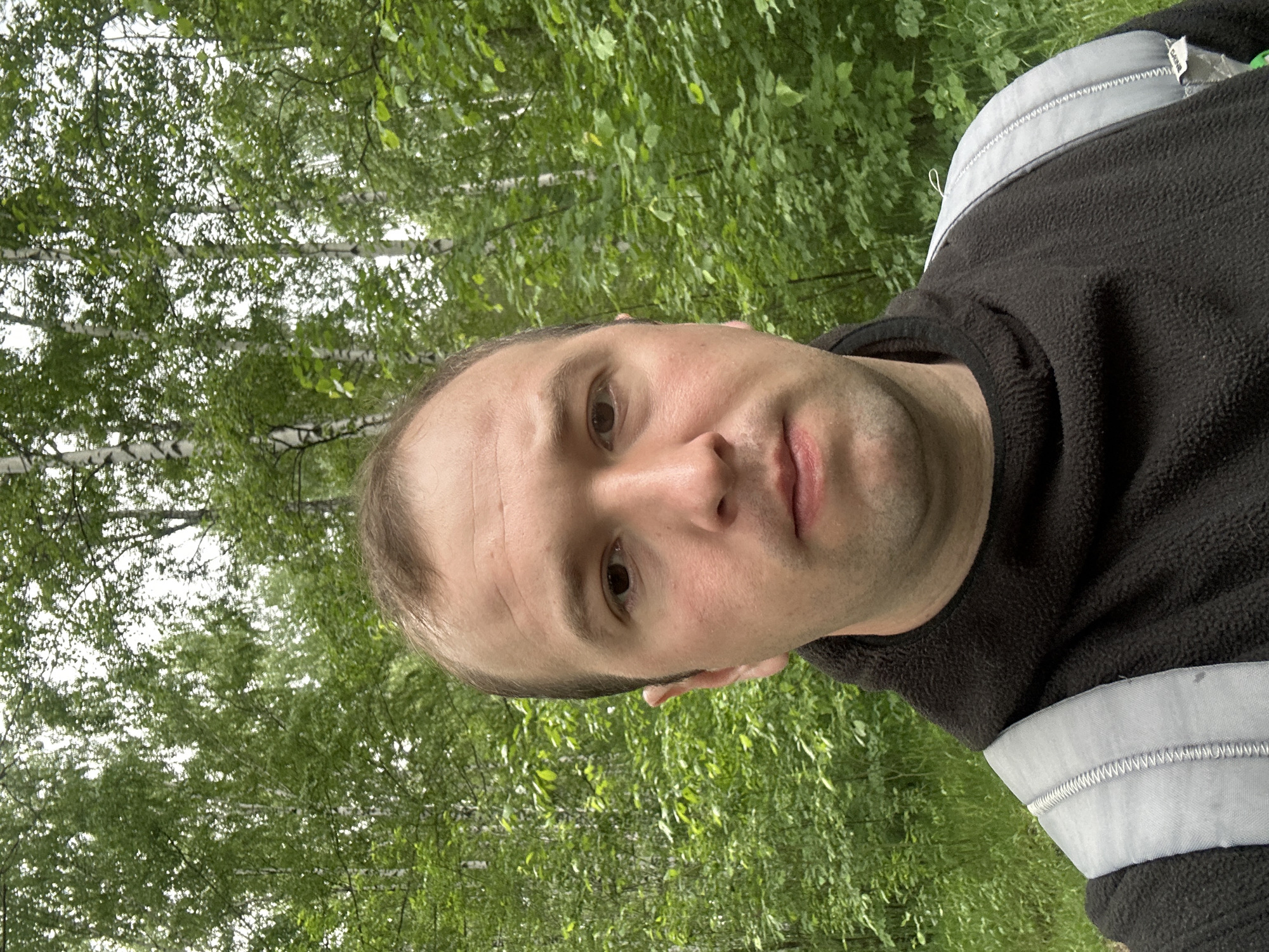 A self-portrait shows a man outdoors, looking directly into the camera with a neutral expression. He appears to be in his late 20s or 30s, with light skin and short brown hair. He is wearing a dark, possibly black or dark grey, fleece-like top. A notable detail on his clothing is a light grey or silver strip with visible zigzag stitching on the upper arm/shoulder area, which might be a reflective element or decorative trim. A tag on his clothing, partially visible in one of the crops, displays the text "SABNIB".
The background is a dense forest or wooded area, filled with vibrant green foliage and the distinctive white bark of birch trees, suggesting a setting in Chaikovsky, Russia. The image is captured during daylight hours, likely under overcast conditions, as indicated by the soft, diffused lighting and the lack of harsh shadows. The lush greenery points to a spring or summer season. The original image is oriented 90 degrees clockwise.