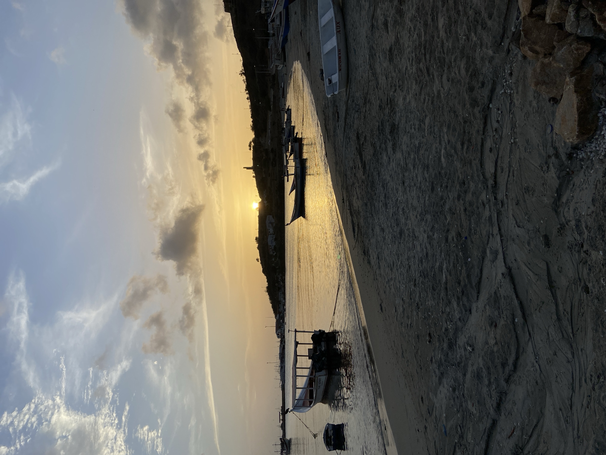 This is a vertical, eye-level, medium shot of a beach in Juan Griego, Venezuela, during sunset. The sky is a gradient of pale blue at the top, fading into a soft yellow and orange hue near the horizon. Wispy clouds are scattered across the sky. The sun, a bright yellow orb, is visible setting behind a dark, hilly coastline dotted with buildings and streetlights.
In the foreground, a sandy beach stretches from the bottom of the frame to where the water meets the shore. The sand is a dark gray and shows textures of rippled patterns, possibly from the tide or wind. Some small rocks and pebbles are scattered on the sand, and a few larger rocks are piled in the bottom right corner.
The ocean reflects the warm colors of the sunset, creating a shimmering golden surface. Several small to medium-sized fishing boats are moored in the water. One boat, a light-colored vessel with a canopy and two outboard motors, is tied to the shore with a rope. Another smaller, dark boat is floating nearby. Further out in the water, more boats are visible, some appearing as silhouettes against the sunset. A few birds are perched on the boats and the shoreline. The coastline curves to the right, with buildings visible on the slope of the hill.