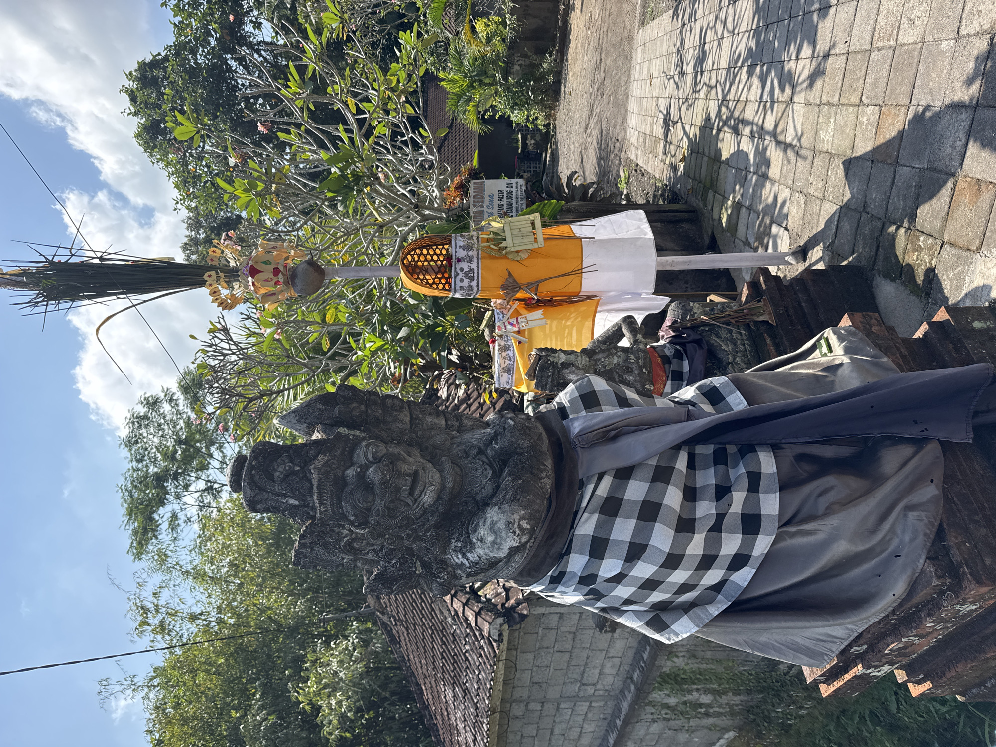 In the Ubud District of Indonesia, a stone statue of a gargoyle-like figure stands adorned with a black and white checkered sarong and a dark grey sash. The statue is positioned on a brick base. To the right of the statue, a tall pole is decorated with traditional Balinese offerings, including yellow and white fabric, woven baskets, and dried leaves. Behind the statue, a tiled roof structure and a stone wall are visible. Lush green trees and vegetation surround the scene, with a paved pathway extending into the background. The sky is bright blue with scattered white clouds, indicating a sunny day. A sign with Indonesian text is partially visible in the background.