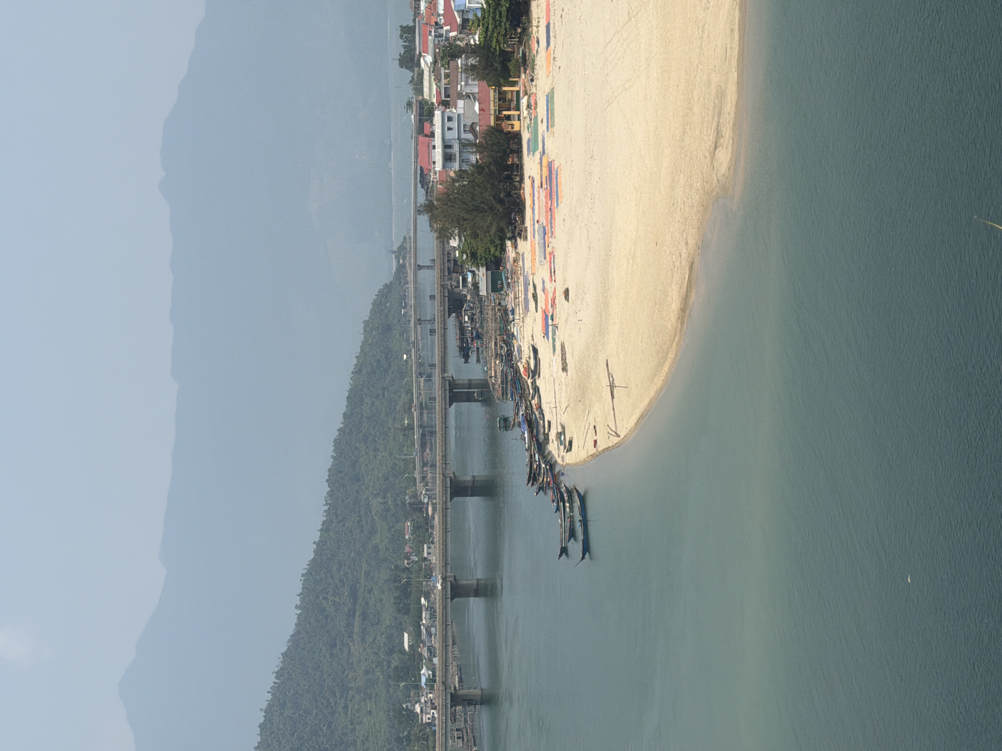 Here's a description of the image:
The image shows an aerial view of a section of the city of Huế, Vietnam, focusing on a coastal area. A bridge spans a river that flows into the sea. A long sandy beach stretches along the coastline, with boats docked at the river's edge.
In the background, mountainous terrain is visible, partially obscured by mist or haze. Buildings are visible along the coast and on the opposite side of the river, close to the bridge. Some colorful fabrics are arranged on the beach.