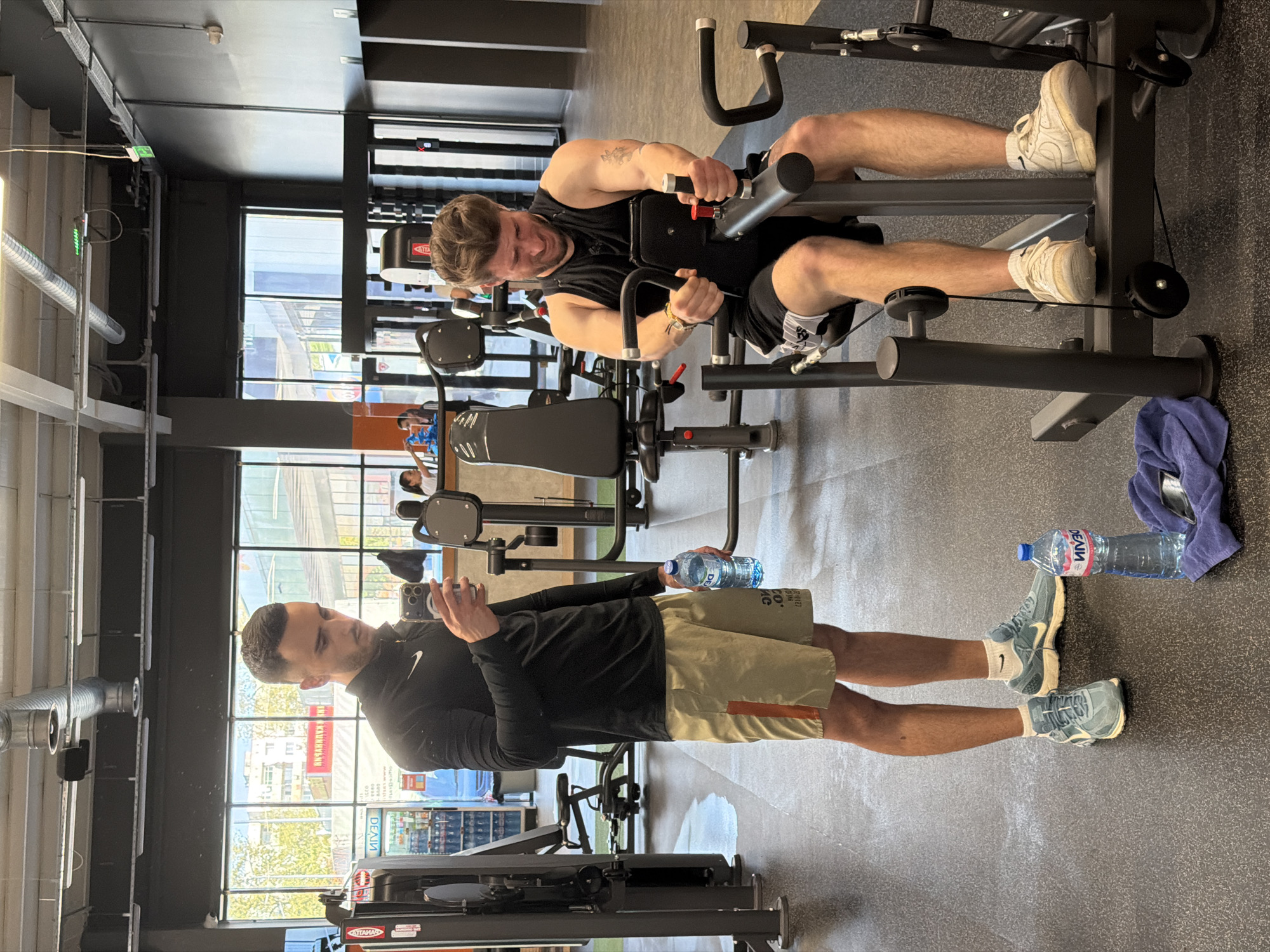 This image captures an indoor scene within a modern gym or fitness center during daytime, characterized by ample natural light streaming through large windows. The interior features a dark ceiling, exposed silver ventilation ducts, and dark gray flooring, suggesting an industrial aesthetic.
Two men are present. In the background, one man with light brown hair and a beard is actively engaged in exercising on a black weight machine, likely a chest press or rowing machine. He is wearing a black sleeveless top and black shorts, with a visible tattoo on his left arm. His facial expression indicates effort as he grips the machine's handles. He wears white athletic shoes and white socks.
In the foreground, another man stands casually, holding a smartphone in his right hand as if taking a photo or video. He holds a clear plastic water bottle in his left hand. This man has dark, neatly styled hair and is dressed in a long-sleeved black Nike shirt and khaki shorts with an orange stripe. He is wearing blue/gray athletic shoes with white Nike-branded socks.
Notable details include a purple towel and another water bottle resting on the floor near the exercising man. The brand "ДЕВИН" (Devin), a well-known Bulgarian mineral water, is clearly visible on the water bottle held by the standing man, and another bottle on the floor. Reflections in the large windows show blurry external buildings and what appear to be Cyrillic letters on signs, further supporting the location context of Plovdiv, Bulgaria. The Nike swoosh logo is prominent on the standing man's shirt and socks.
