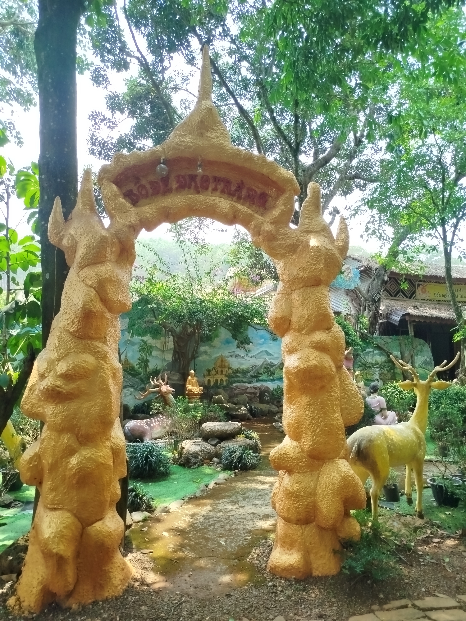 This media file captures a serene outdoor scene, likely an entrance or garden feature in Nam Ban, Vietnam. The primary subject is a large, golden, ornate archway resembling a stylized tree or antlers, serving as a gateway. Its surface has a textured, rough appearance.
Through the archway, a landscaped area is visible, featuring various statues and a painted mural in the background. Two golden deer statues are prominent: one standing gracefully on the right, and another spotted deer lying down further inside to the left. A golden Buddha statue is also visible, along with other smaller figures or statues, nestled among green foliage and rocks. The mural behind these figures depicts a landscape with trees, temples, and possibly a body of water.
In the far background, behind the left side of the archway, a building with a thatched roof and intricate patterns is discernible. A sign on this building shows partially visible Vietnamese text, including phrases like "CÁ SA ĐI" and "Đến ngày". Lush green trees frame the scene, with bright daylight filtering through their leaves, suggesting a clear, sunny day. Shadows are cast on the ground, indicating the time is likely morning or afternoon.
The text inscribed on the golden archway appears reversed or mirrored, reading something like "BODE BHOTRAA" from left to right as seen in the image, possibly stylized or intended to be read from the opposite side. The overall atmosphere is peaceful and decorative, characteristic of a spiritual garden or a tourist attraction.