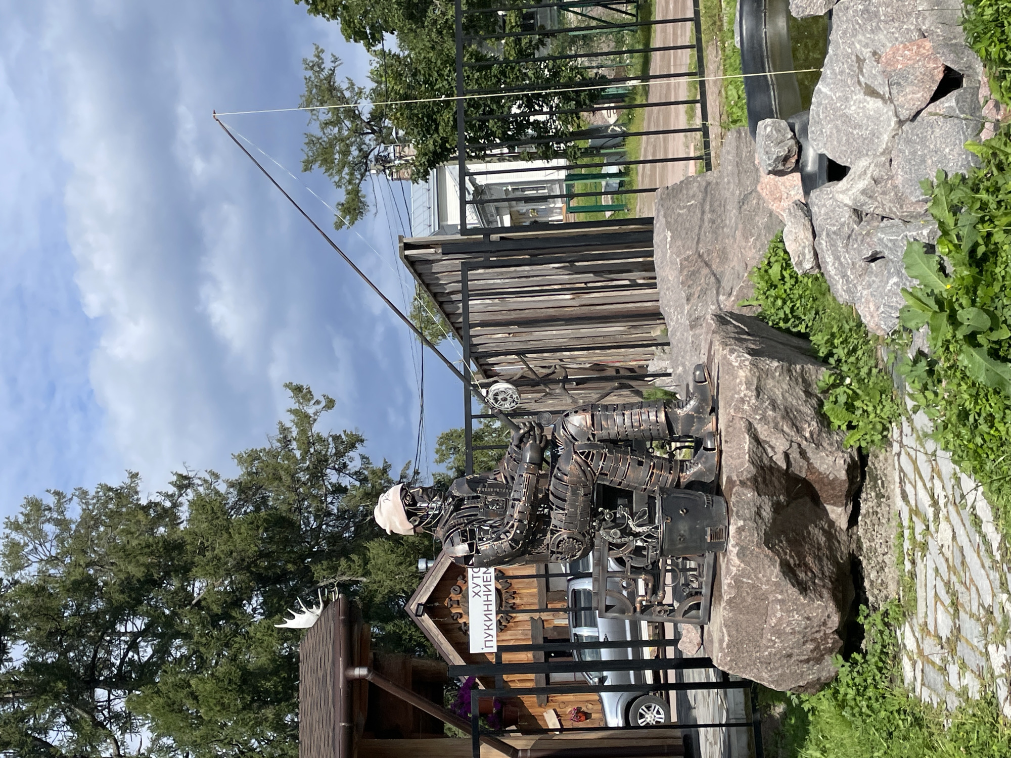 In Berozovo, Russia, a metal sculpture of a fisherman sits on a large rock, holding a fishing rod. The sculpture, constructed from various metal parts, depicts a figure wearing a hat and fishing gear. In the background, a wooden building with a sign reads "ХУТО ПУКИННИЕМ". A silver car is parked behind a black metal fence, and trees and a cloudy sky complete the outdoor scene.
