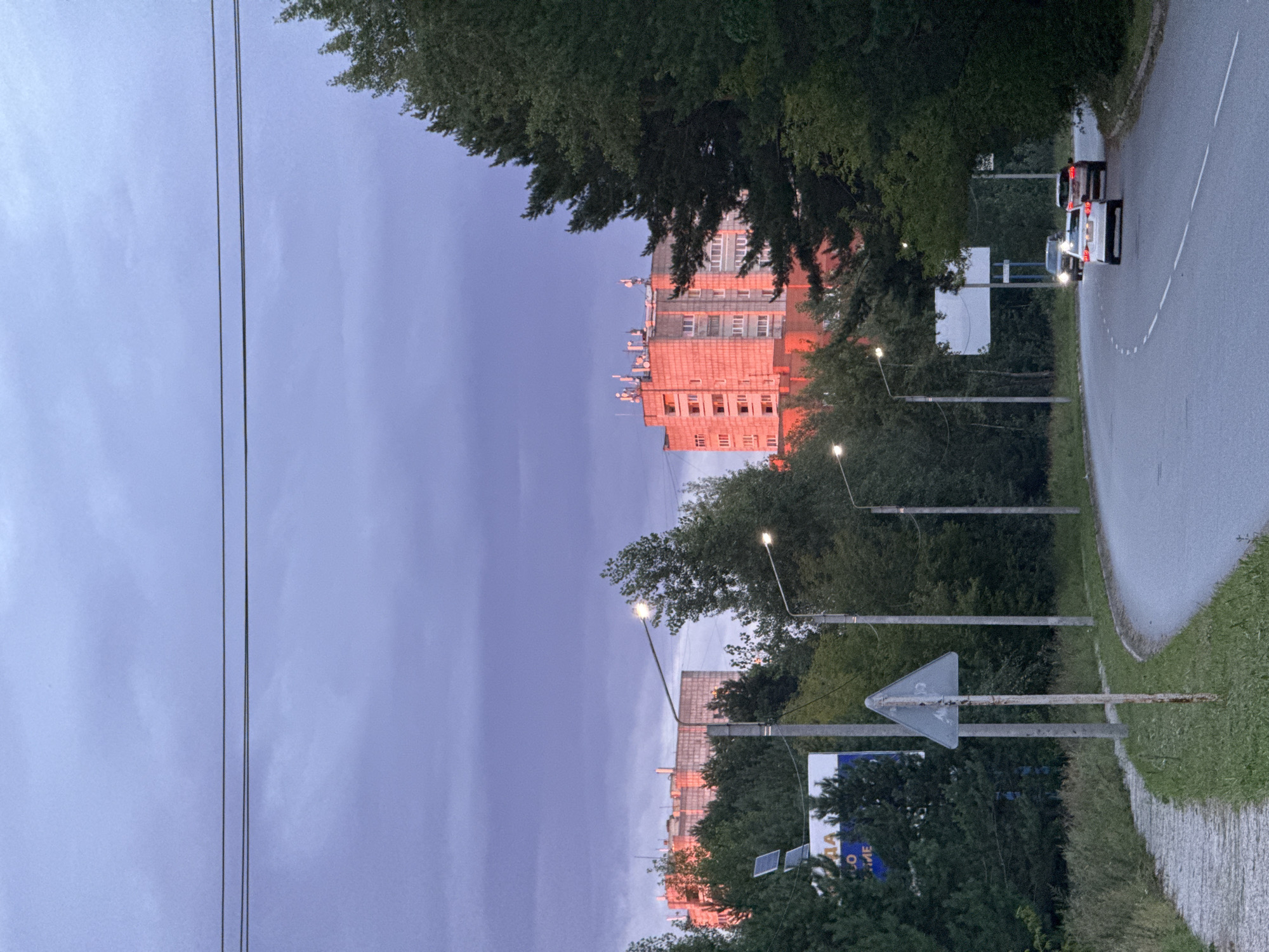 An outdoor, eye-level, wide shot captures a curved asphalt road winding through a green, overgrown area with a sidewalk on the left. In the background, a tall, multi-story building with a pinkish-orange facade illuminated by the setting sun stands amidst dense trees. Several vehicles, including a white sedan and two SUVs, are driving on the road. Streetlights are visible along the roadside, and power lines stretch across the dusky, purple-grey sky. A triangular road sign is planted on the side of the road. The scene appears to be in a suburban or semi-urban environment.