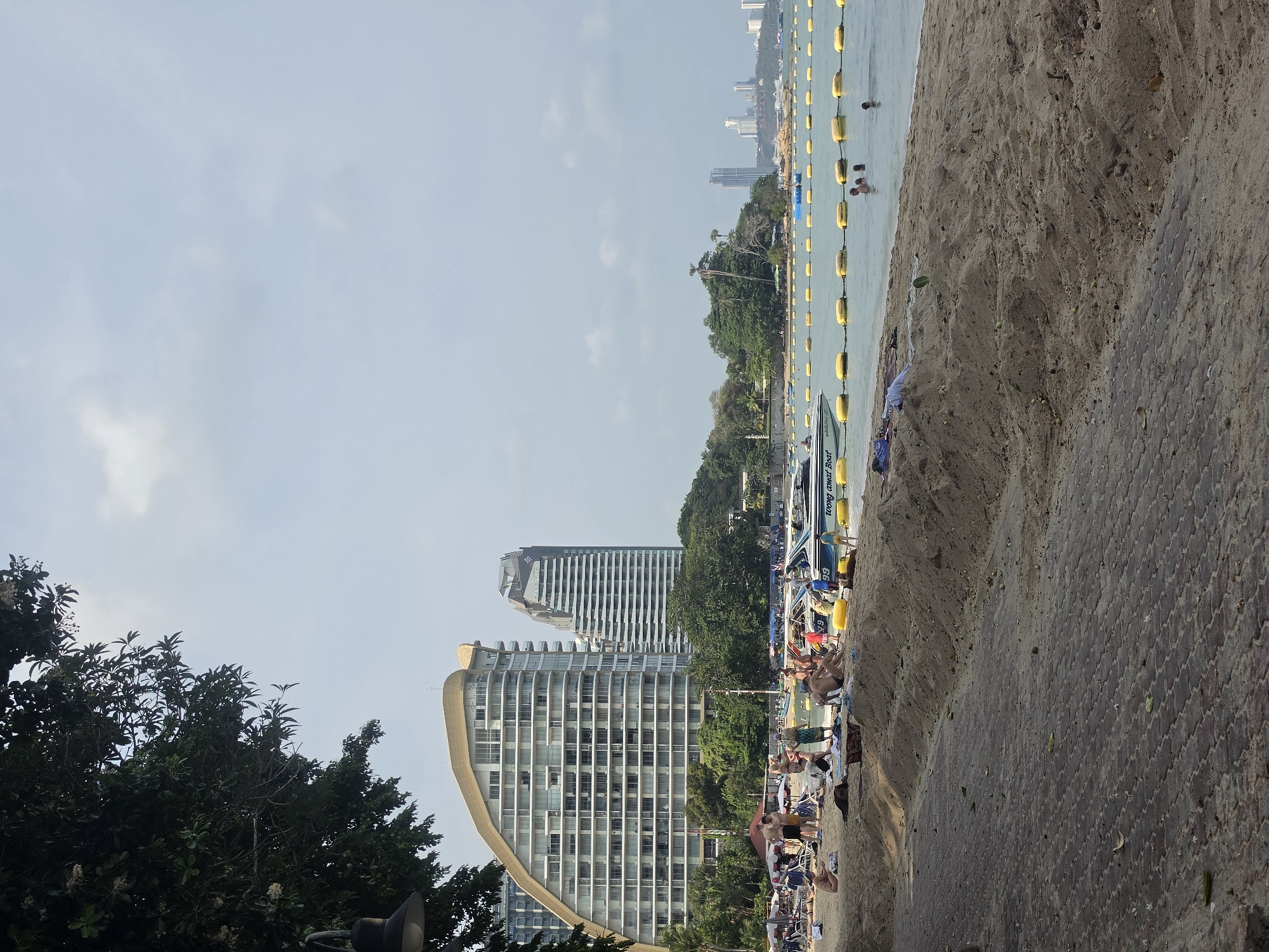 This image depicts a sunny daytime scene at a beach in Pattaya City, Thailand, with an urban coastline backdrop.
In the foreground, a textured, paved walkway runs parallel to a strip of sandy beach. Several people are visible relaxing on the sand, some near small boats. One boat clearly displays the text "Wong amat Boat" along with the numbers "69" and "6A" on its hull.
The midground features the calm, light blue ocean. A long line of yellow buoys extends across the water, likely delineating a swimming zone or boat path. A few individuals are seen swimming or wading in the sea. Beyond the immediate beach and water, dense green trees and tropical foliage line the coast.
The background is dominated by the skyline of Pattaya City, showcasing numerous modern high-rise buildings and skyscrapers, indicating a developed urban area. The sky above is a clear, pale blue with only a few wispy clouds, suggesting pleasant weather for beach activities. The overall scene conveys a relaxed, leisure atmosphere typical of a popular coastal resort.