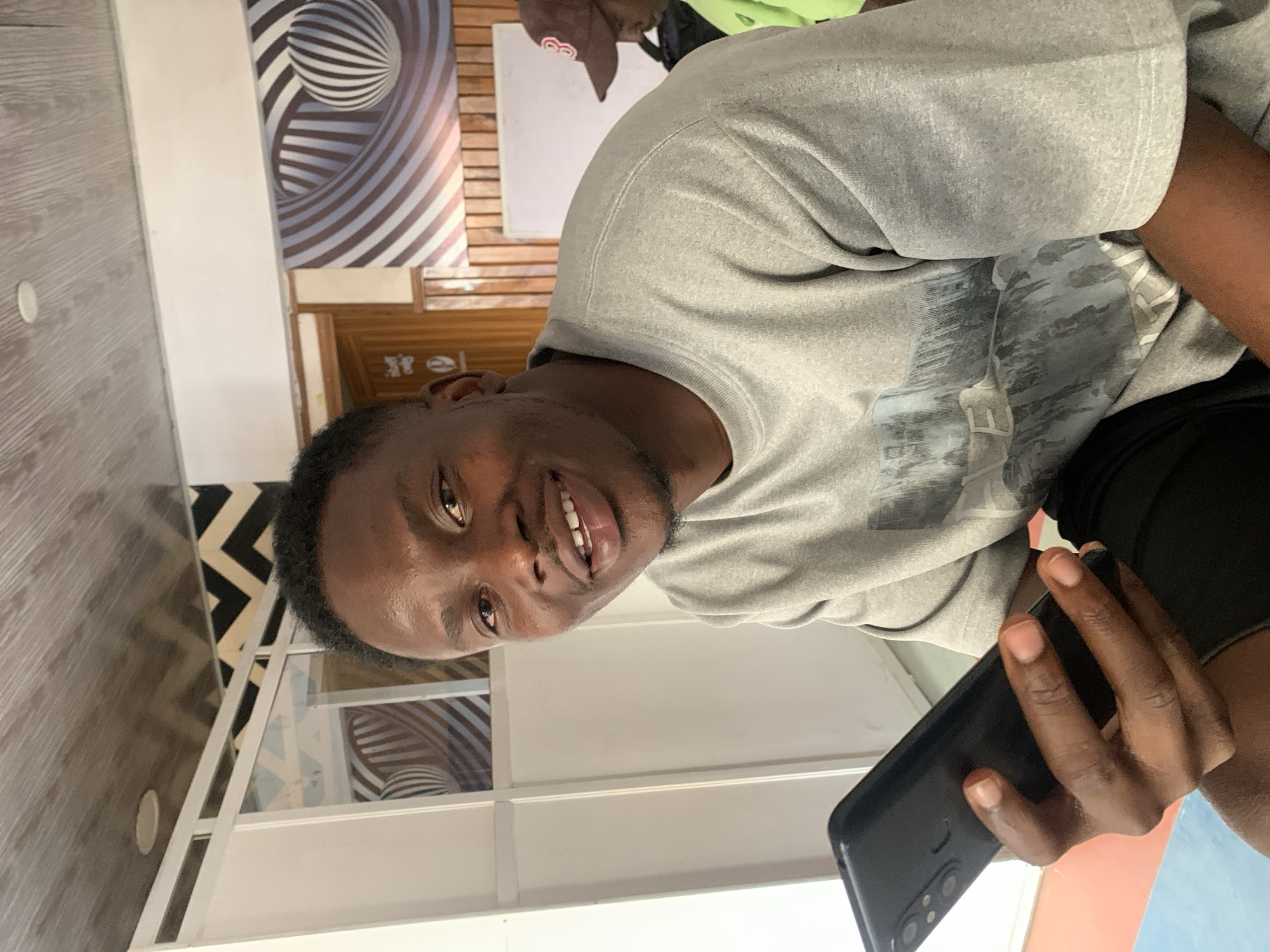 A young Black man is captured in a close-up, chest-up portrait shot, looking directly at the camera with a broad, friendly smile that reveals his teeth. He has short, dark hair and light stubble. He is wearing a casual grey short-sleeved t-shirt featuring a subtle graphic design in blues and greys across the chest, and dark shorts or trousers. In his right hand, he holds a black smartphone with multiple camera lenses visible on its back.
The setting appears to be indoors, with bright, even lighting. The background features a mix of textures and patterns. To the man's left, there is a wall with a light wooden grain pattern. Behind him and to his right, a striking black and white optical illusion artwork, possibly a poster or wallpaper, depicting swirling stripes and a sphere, adorns a wall. Below this, another wall section has a wooden or brick-like texture. Further into the background, part of another person wearing a brown cap and a green top is visible, and a white board can be seen. On the floor to the left, a black and white chevron or zig-zag patterned rug is visible. White room dividers or window frames also contribute to the indoor office or common area aesthetic, typical of a professional or communal space in a city like Jos, Nigeria. No discernable text is visible.