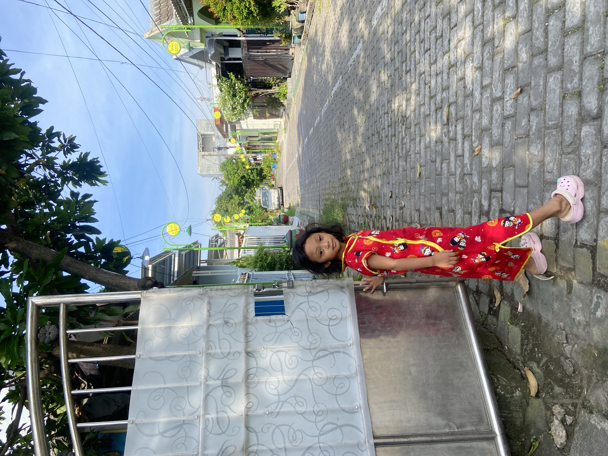A young girl wearing a red Chinese-style dress and pink clogs stands next to a metallic sliding gate on a cobblestone street in Gelam, Indonesia. The street is lined with houses and green street lamps. Trees and power lines are visible against a blue sky.