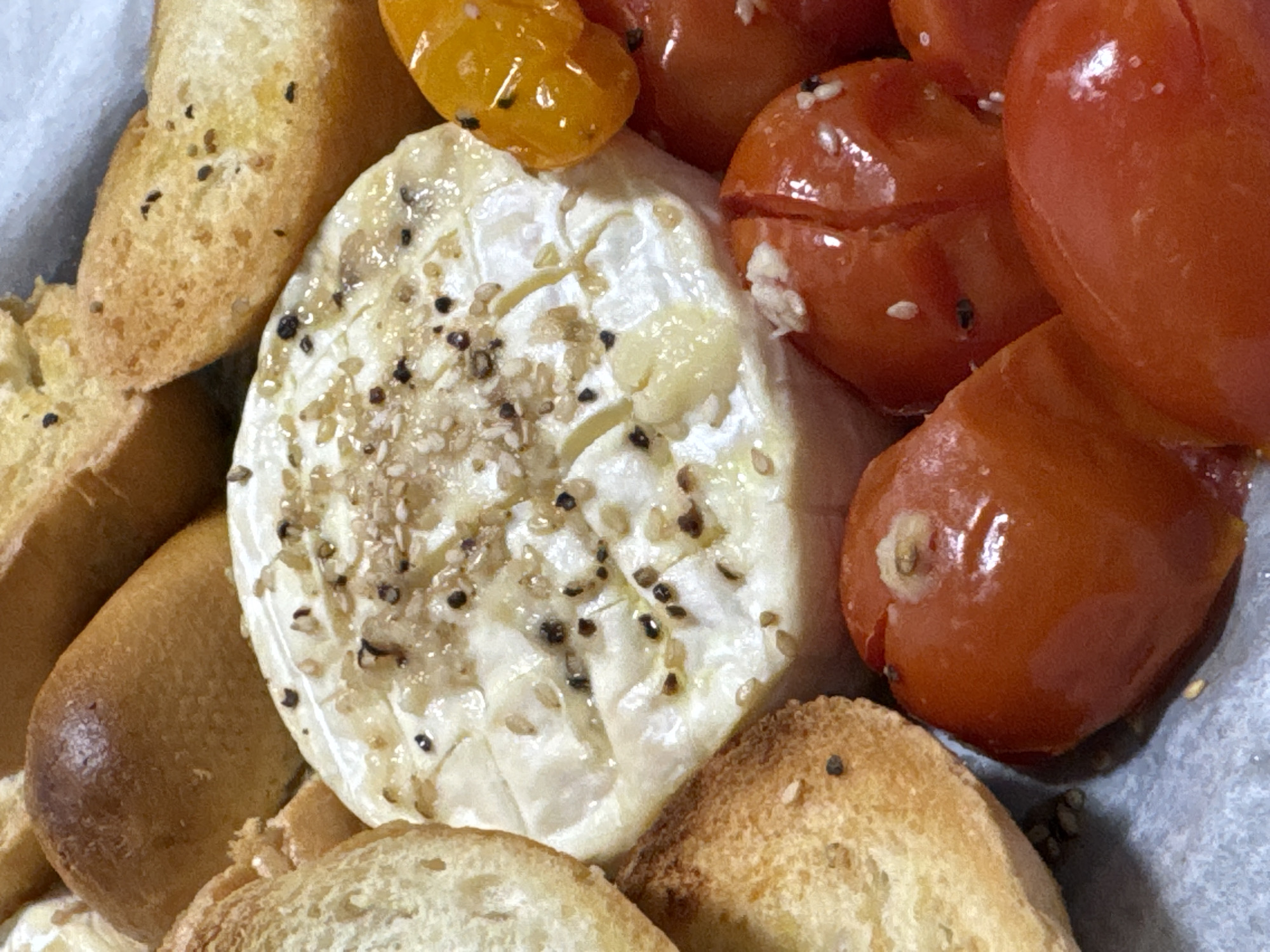 The image shows a close-up of food arranged on a surface covered with parchment paper. There are slices of toasted bread, a round cheese with what appears to be oil and seasonings on top, and a pile of red and yellow cherry tomatoes. The location in Ramat Gan, Israel is not apparent from the image.