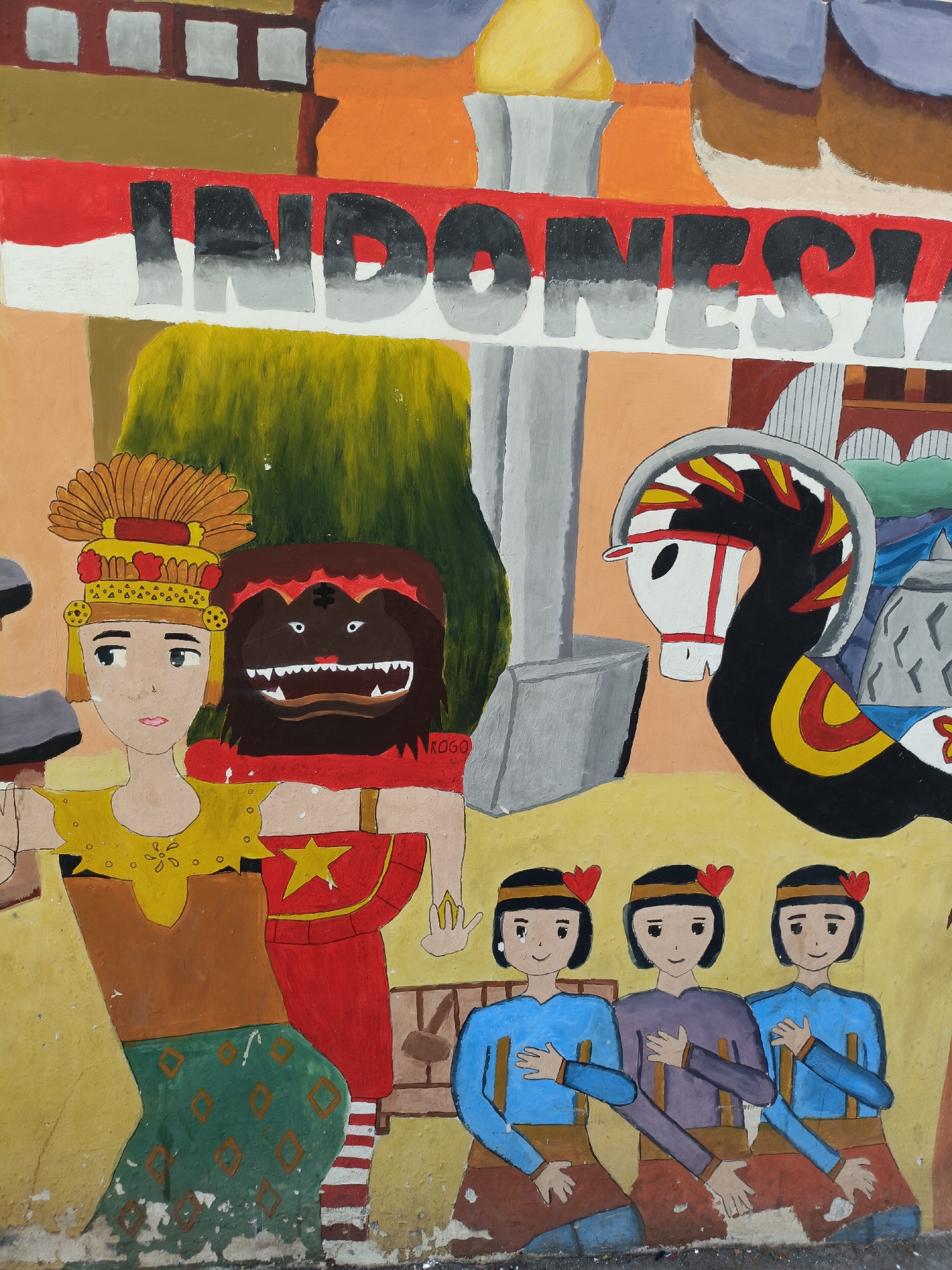 This is a colorful, folk-art style mural painted on a weathered wall, likely in Kediri City, Indonesia. The mural depicts various cultural elements. Dominating the upper portion of the mural is the word "INDONESIA" painted in large, grey, 3D-style letters across a red and white striped background, representing the Indonesian flag. Below this, to the left, stands a person in traditional attire, including an elaborate headdress. Next to them, on the left, is a figure with a brown, furry face and sharp teeth, resembling a mask or a mythical creature. To the right of these figures is a stylized representation of a horse head, possibly part of a larger depiction of a horse or a parade float, adorned with yellow and red accents. In the lower section of the mural, three figures, presumably young women, are seated, wearing blue tops and brown skirts, with red heart-shaped decorations in their hair. The overall scene appears to celebrate Indonesian culture and national identity. The wall shows signs of age and wear, with peeling paint and visible textures.