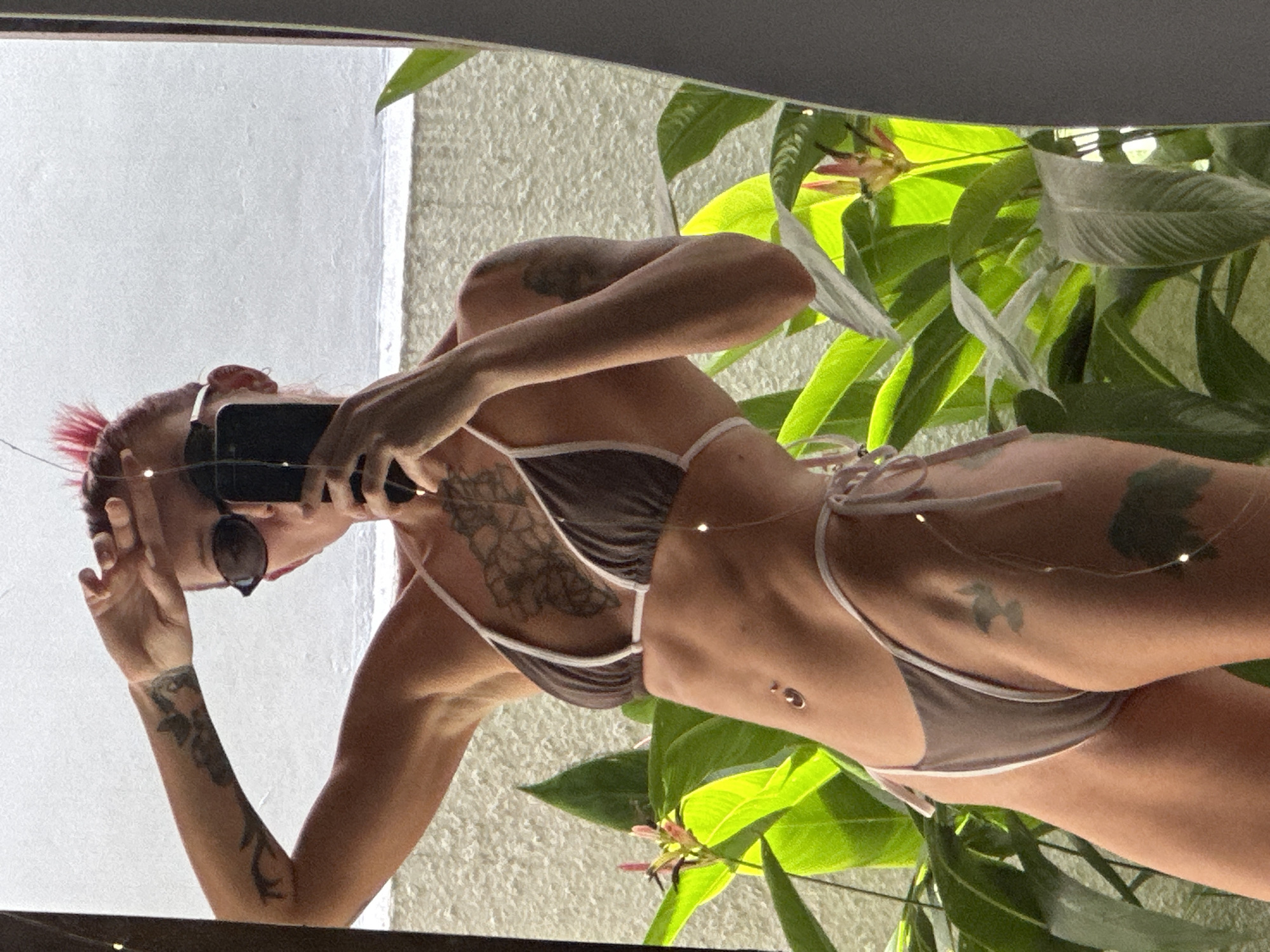 A woman with pink-red hair and tattoos stands indoors, taking a picture of herself in a mirror with her phone. She is wearing a brown bikini with white trim. The reflection shows outdoor greenery, possibly in Dalung, Indonesia, due to the tropical foliage visible. The background features a white textured wall.