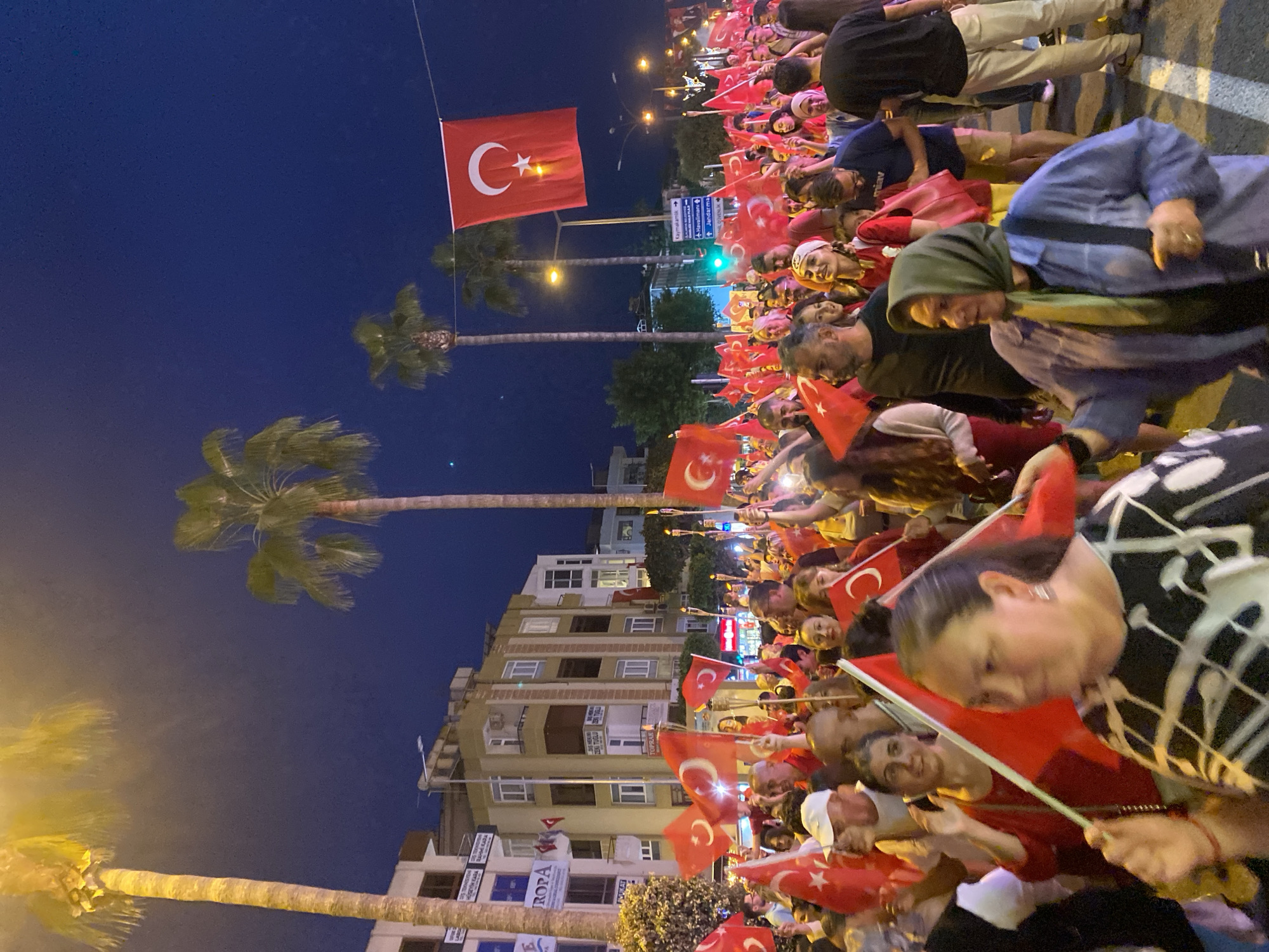 A crowd of people waving Turkish flags are gathered on a street in Alanya, Turkey. The crowd includes women and men, some wearing headscarves. Buildings line the street, with signs visible on the storefronts. Palm trees are visible in the background. A large Turkish flag hangs from a wire above the street. The sky is dark blue, suggesting it is night or dusk. A traffic light is visible in the distance.