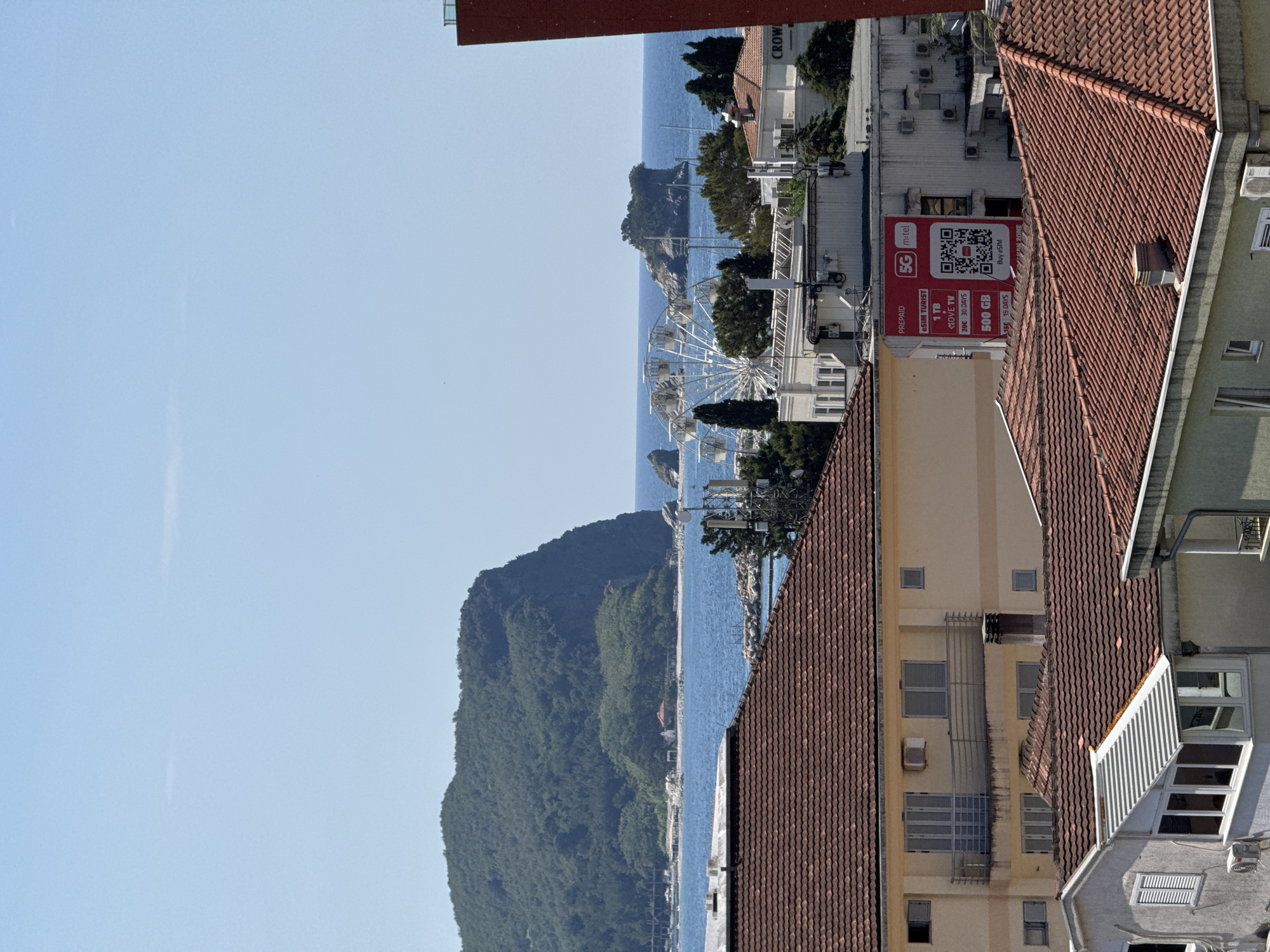 This is a medium-shot, eye-level, outdoor photograph taken on a clear day, with a bright blue sky dominating the upper half of the frame. The scene depicts a coastal view with a mix of natural landscape and urban structures.
In the foreground, the tops of several buildings with terracotta tiled roofs are visible, suggesting a residential or mixed-use area. These buildings have light-colored facades and various window styles. Some have balconies with metal railings, and one building has an air conditioning unit. Chimneys also protrude from some of the roofs.
The middle ground features a more distant row of buildings, including a larger white building with multiple windows and visible air conditioning units on its exterior. Attached to this building is a large red advertisement for "5G m-tel" offering prepaid eSIM tourist plans with varying data amounts and durations, along with a QR code. To the right, a dark red, vertical structure, likely part of another building, partially obscures the view. Beyond these buildings, a white Ferris wheel stands out against the blue sea, suggesting a recreational area.
The background is dominated by a lush green, forested island or peninsula jutting out into the calm, blue sea. The island has steep, rocky cliffs on its seaward side. Smaller rocky islets are scattered around it. The horizon line is low, emphasizing the expanse of the sea and sky.
The overall impression is a sunny day in a coastal town or city, where natural beauty meets modern infrastructure. There are no people visible in the image.