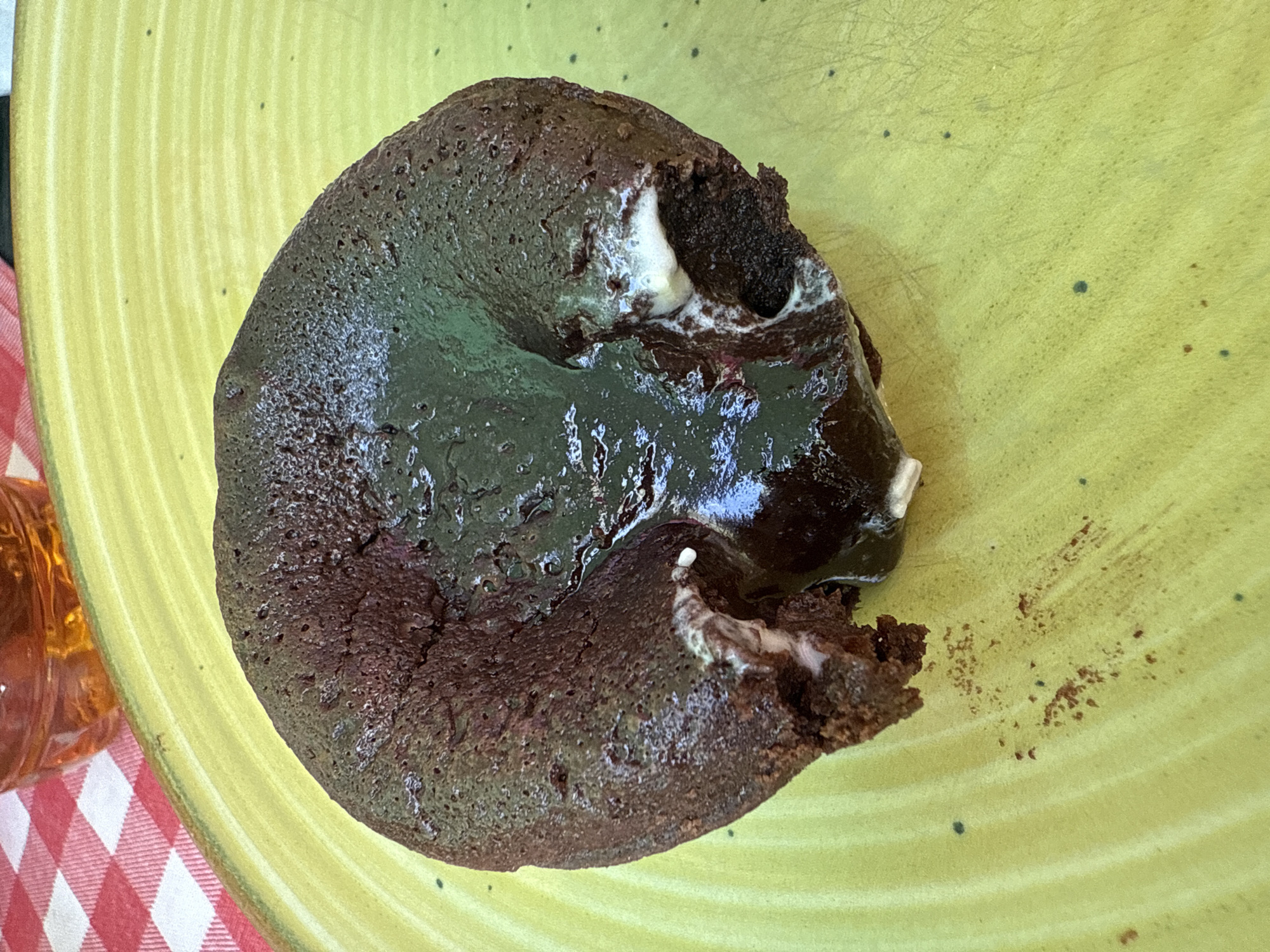A single lava cake sits on a light green plate. The cake is dark brown with a molten chocolate center and a small amount of white cream or frosting. Part of the cake has been broken off. The plate has small, dark spots. A glass with an orange liquid is visible in the lower left corner of the image, next to a red and white checkered surface. The image was taken in Novo Oriahovo, Bulgaria.