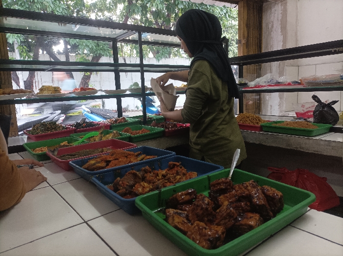 A woman in a hijab stands behind a counter filled with various dishes in green and blue trays. She is reaching into a paper bag. The trays contain a variety of foods with different colors and textures, including vegetables, meat, and fried items. Above the counter are shelves with more dishes and a window showing a street scene. The setting appears to be a small restaurant or food stall.