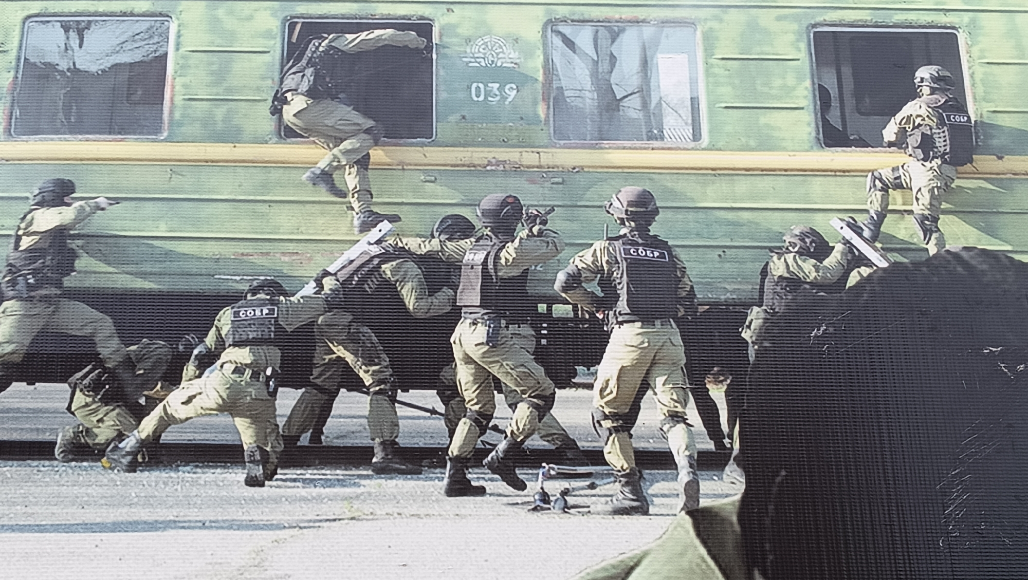 This image captures a dynamic scene depicting what appears to be a special forces unit conducting an assault or training exercise on a green train carriage, likely at a railway station or yard in Tiraspol, Moldova.
Numerous individuals, dressed in tan/khaki tactical combat uniforms, helmets, and protective vests, are actively engaging with the train. Several operators are seen climbing or entering the train through its windows. One figure is seen scaling into an upper-left window, another is mid-entry into an upper-right window, and at least two more are positioned around the windows at a lower level. On the ground next to the train, multiple operators are in various tactical stances, suggesting coordinated movement and action. One operator in the lower-center appears to be wielding a breaching tool, possibly a ram or a shield, directed towards the train's side.
The train itself is olive green with a yellow stripe running along its side. Some of its windows are open or appear to be broken, facilitating entry. On the side of the train, the numbers "039" are clearly visible, along with an indistinct emblem above them. A prominent detail is the text "СОБР" (SOBR, the Russian acronym for Special Rapid Response Unit) clearly visible on the back of several operators' tactical vests, identifying the unit involved.
The scene is illuminated by bright daylight, casting clear shadows, suggesting a sunny day. The overall image has a somewhat grainy texture with distinct horizontal lines, characteristic of certain photographic reproductions. The activity conveys a sense of urgency and precision in a high-stakes scenario.