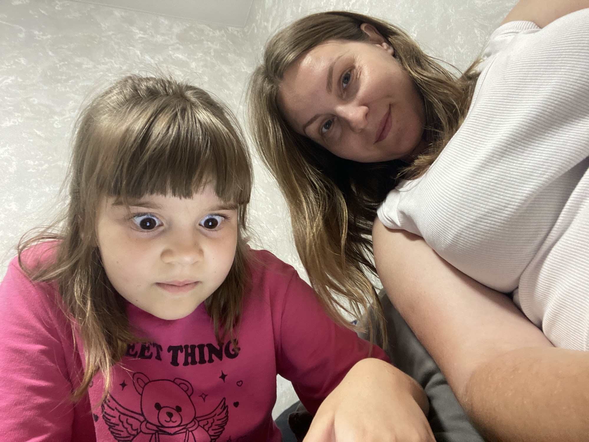 A selfie features a young girl with wide eyes and light brown hair, bangs, and a pink long-sleeved shirt with a graphic of a bear with wings and the text "SWEET THING" printed on it. She is positioned on the left side of the frame, looking directly at the camera. To her right and slightly behind her is an adult woman with shoulder-length light brown hair, also looking at the camera with a slight smile and tilted head. She is wearing a white ribbed shirt. The background is a textured light-colored wall. The image appears to be taken indoors, possibly in a residential setting. There is no discernible indication of the specific city of Noviy, Russia, within the image itself.