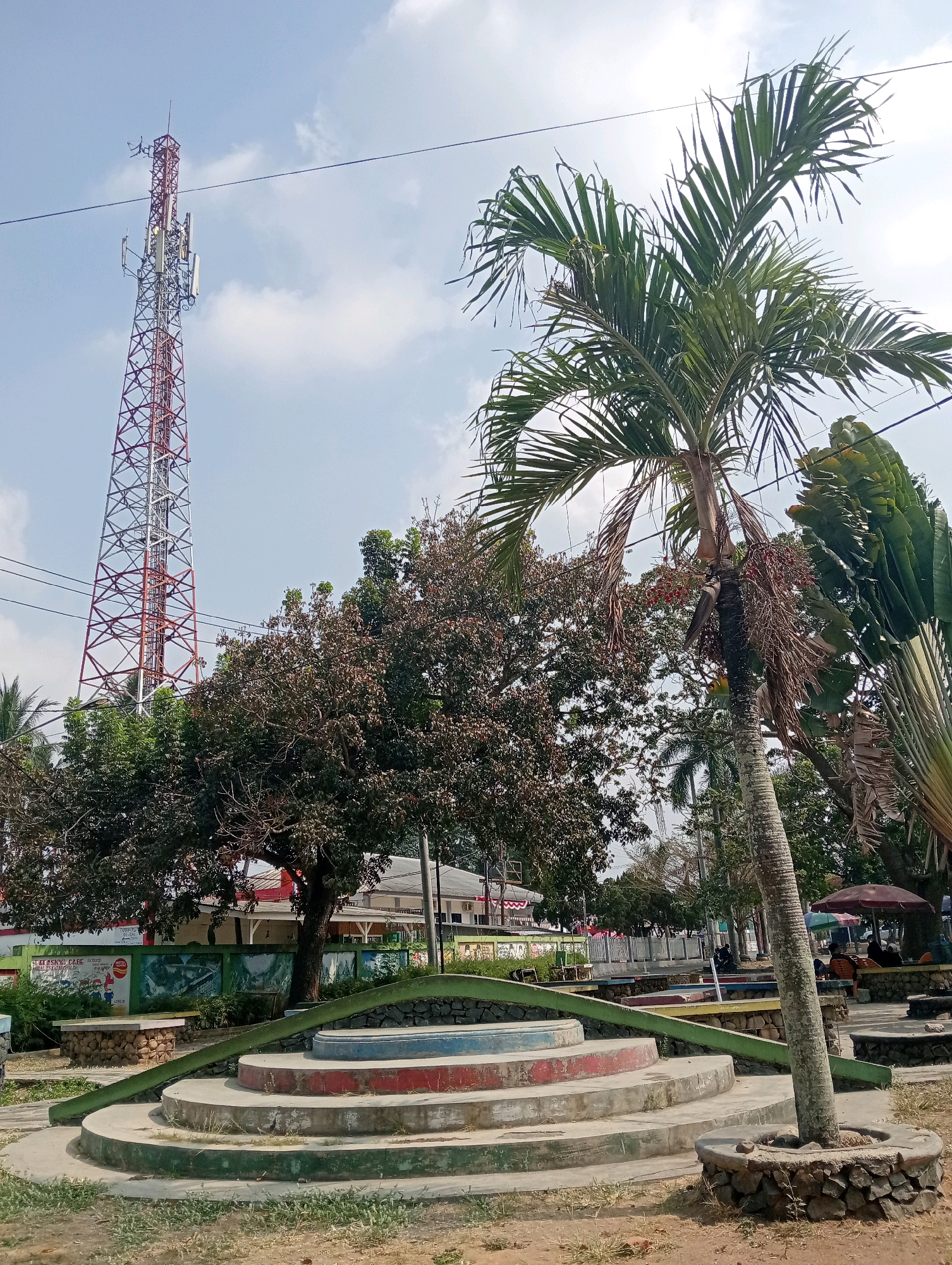 The image shows an outdoor scene in Pringsewu, Indonesia, featuring a large cell tower in the background. In the foreground, there is a multi-tiered concrete structure painted with red and blue accents, surrounded by greenery and a stone border. A palm tree with visible fruit stands to the right of this structure. In the middle ground, several trees, including one with dark foliage, are visible, along with what appears to be a low-lying wall adorned with colorful murals. Beyond the trees, there are buildings, some with awnings, and people sitting under umbrellas at tables. The sky is partly cloudy. Power lines are visible throughout the frame.