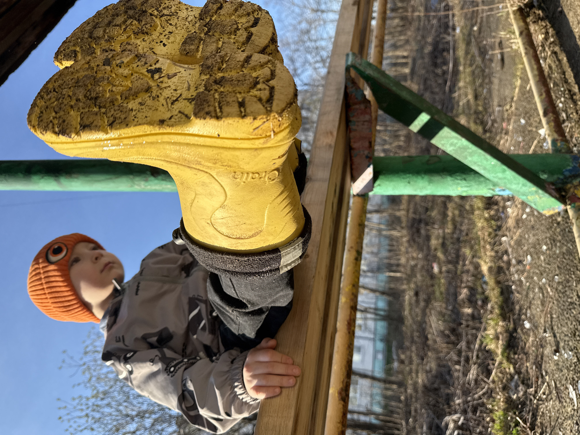 A low-angle shot depicts a child climbing on playground equipment, specifically located in Chaikovsky, Russia. The child, wearing a bright orange beanie with an eye design, a gray patterned jacket, and yellow rain boots, is reaching up a wooden ladder with green metal accents. The ground appears to be covered in dirt and debris. The sky is visible in the background and is light blue. The child's left rain boot is caked with mud. The word "Cralin" is visible on the side of the right rain boot. Some buildings are seen through the branches of the tree in the background.