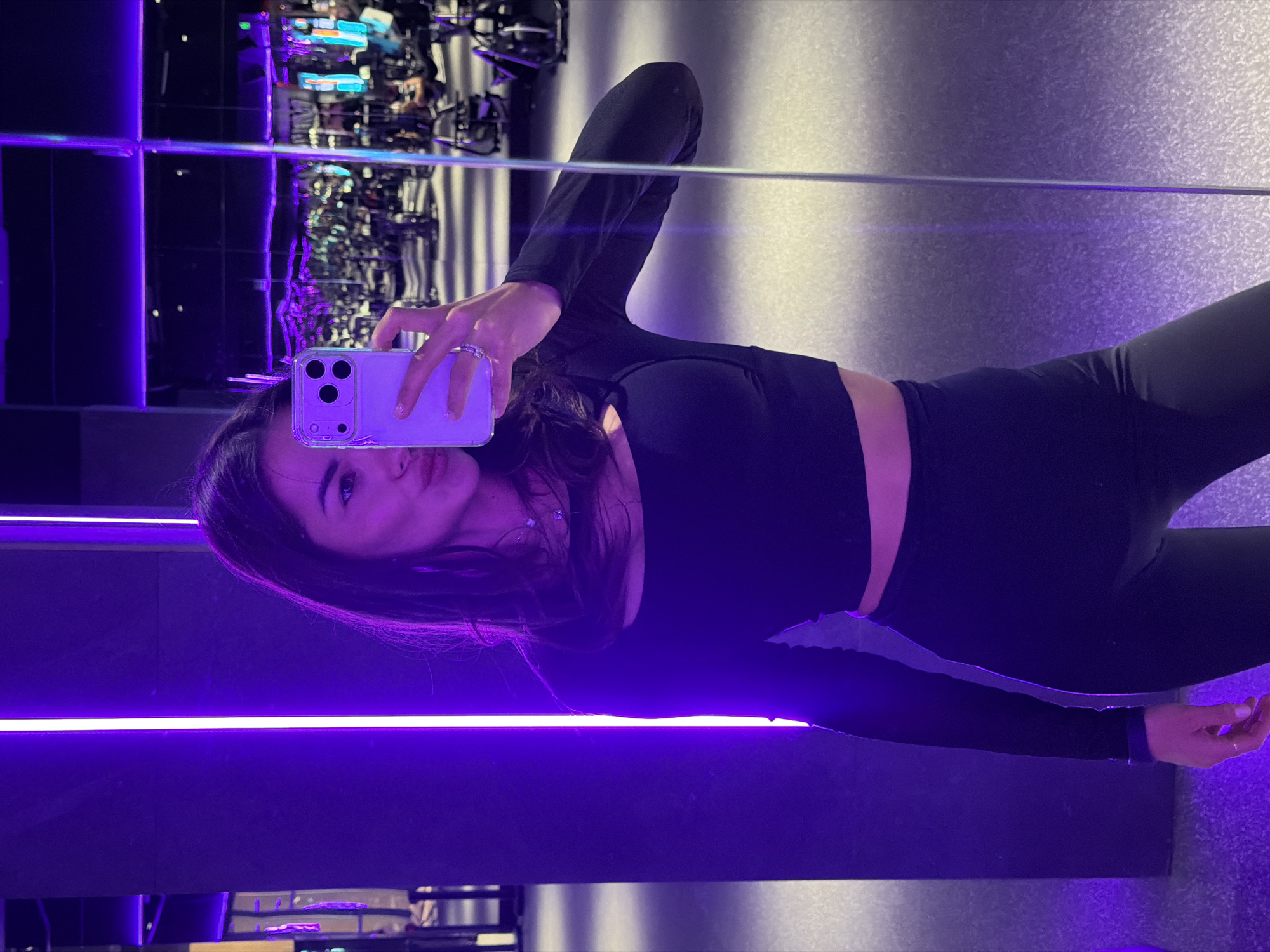A young woman is captured in a mirror selfie within an indoor setting, likely a modern gym or leisure facility in Krasnoyarsk, Russia. She is dressed in a black long-sleeved crop top that exposes her midriff, paired with matching black leggings. Her dark hair is styled down, partially obscuring one shoulder. She holds an iPhone, equipped with a clear case and visible triple camera array, in her right hand, where several rings adorn her fingers.
The environment is illuminated by dramatic purple and blue artificial lighting, creating a vibrant, futuristic, and somewhat moody atmosphere. A prominent horizontal strip of bright purple LED light runs across the lower part of the frame, casting a glow on the surroundings. The mirrored background reflects blurred and abstract shapes of what appear to be exercise equipment or architectural features, further distorted by the colorful lighting. The walls and floor exhibit a dark, textured appearance. The woman's expression is neutral as she looks towards the phone. No discernible text is visible in the image.